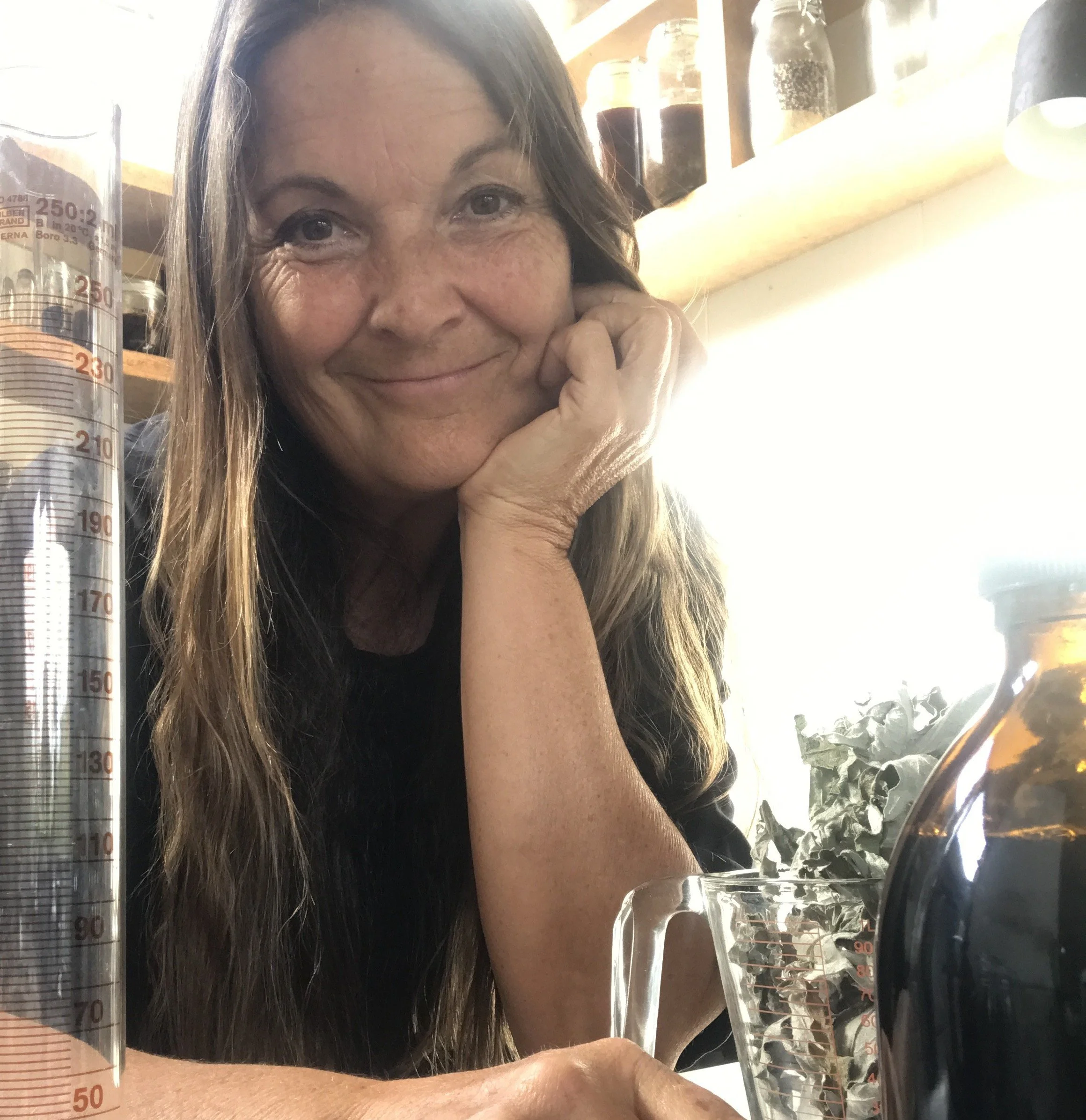 Kerri is smiling, resting her face on her hand, sitting at a table with her herbal medicines.