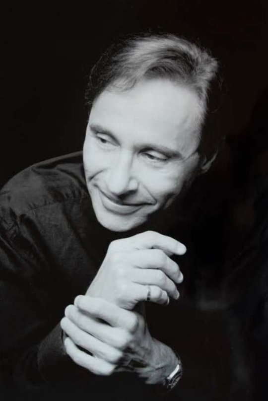 Black and white portrait of a man with short hair smiling, with his hands clasped together, wearing a dark shirt.