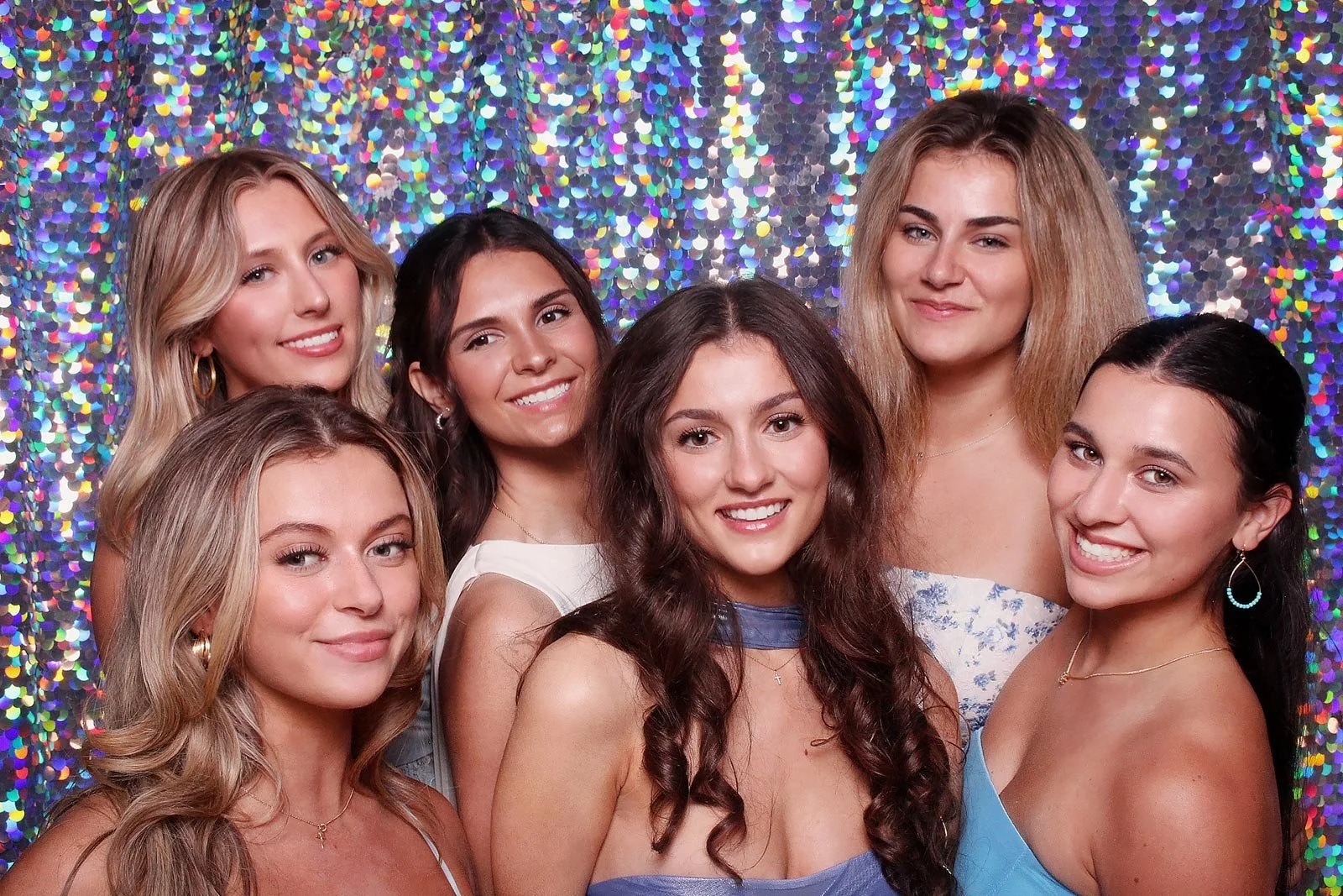 Six young women smiling at a party or event, standing closely together in front of a colorful, sparkly background.