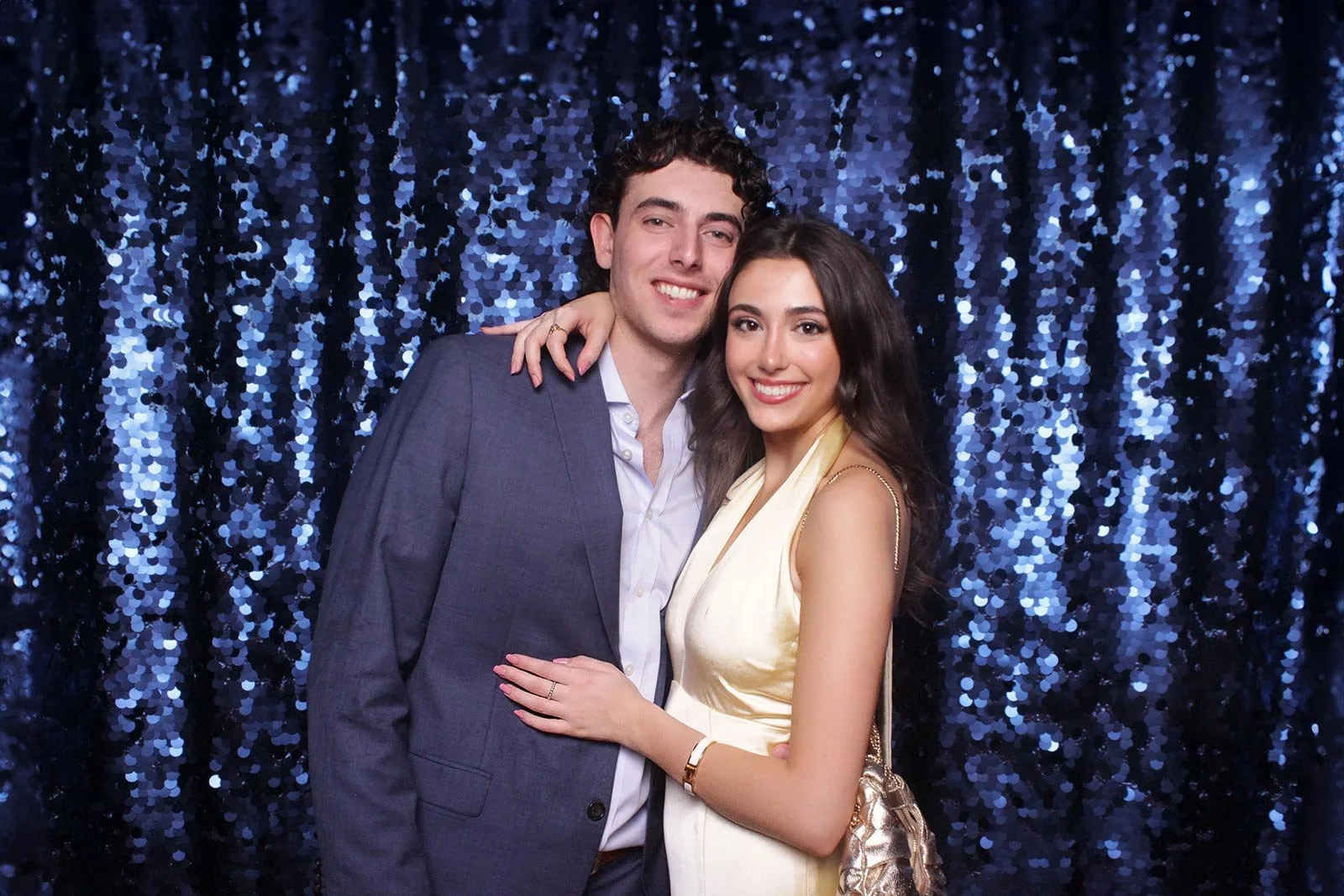 A young couple dressed in formal attire posing together in front of a dark blue sequin backdrop, smiling at the camera.