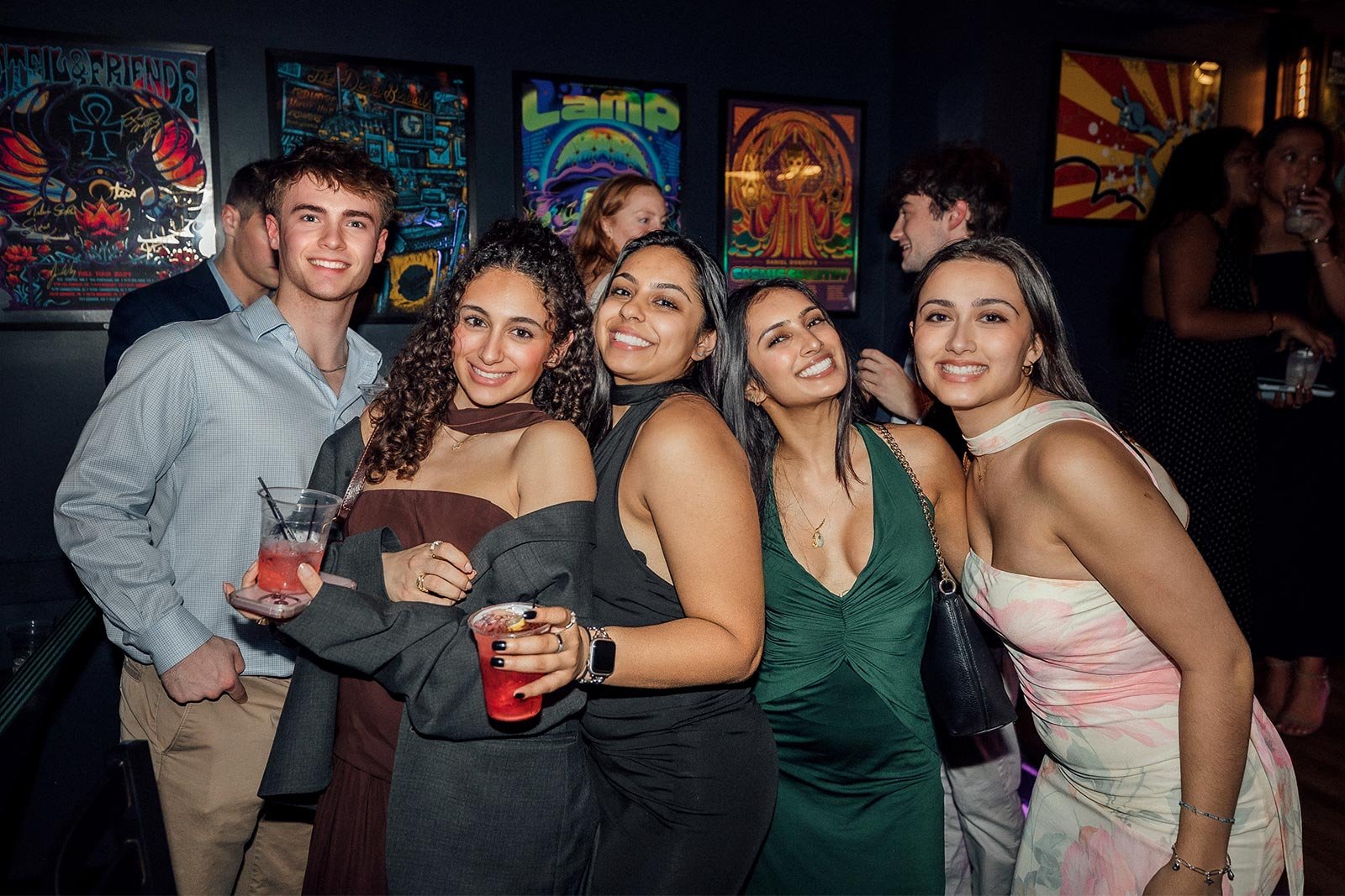 A group of five young adults smiling and posing together at a party, holding drinks in a dimly lit bar with colorful posters on the wall behind them.