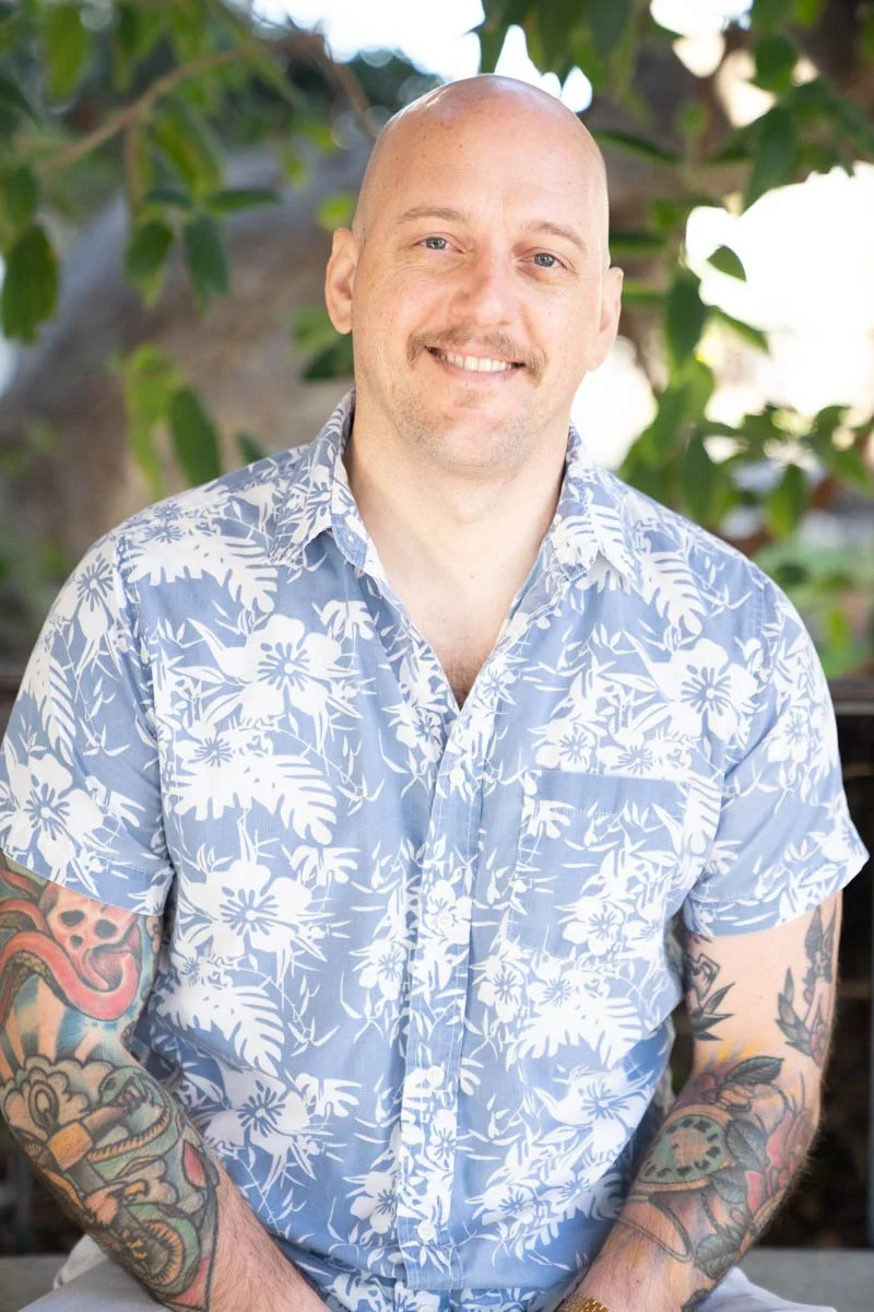 A smiling man with a shaved head and tattoos on his arms, wearing a blue Hawaiian shirt with white floral patterns, stands outdoors with green leaves in the background.