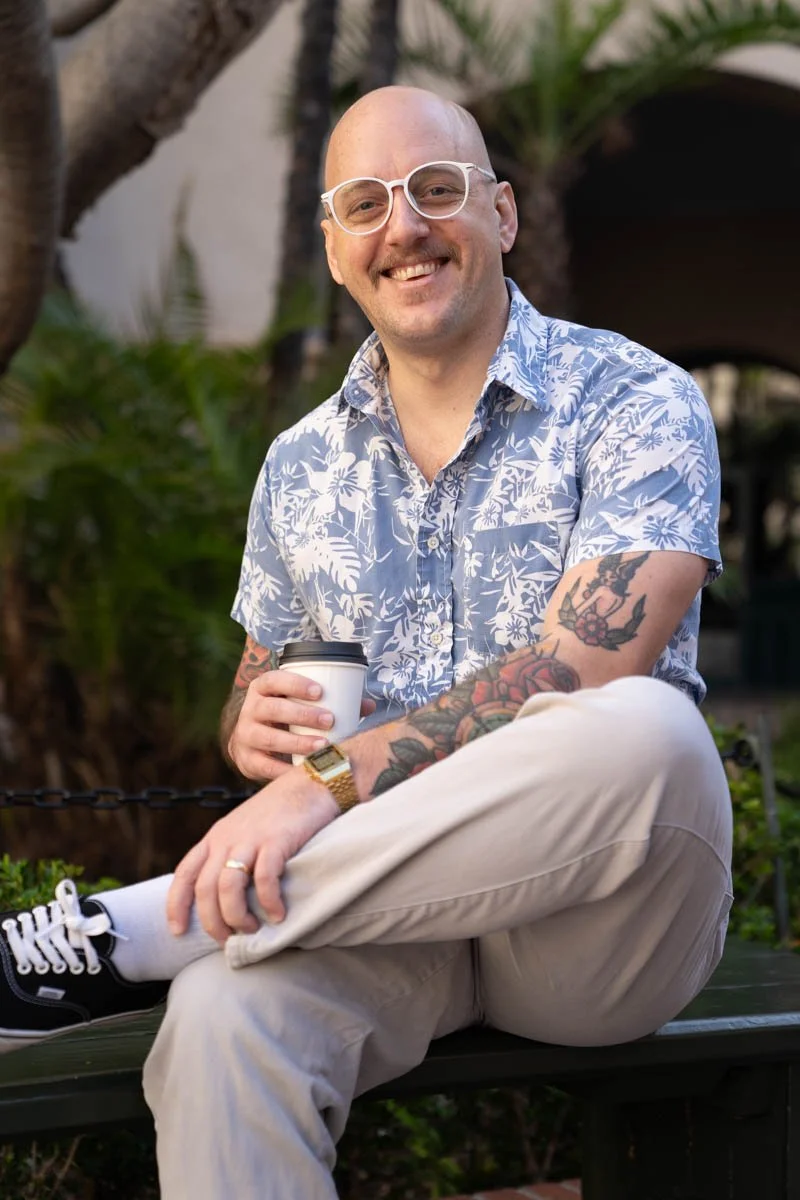 A man with a shaved head, glasses, and tattoos, sitting outdoors on a park bench, holding a coffee cup and smiling at the camera. He is wearing a blue and white floral shirt, beige pants, and black sneakers, with greenery and trees in the background.