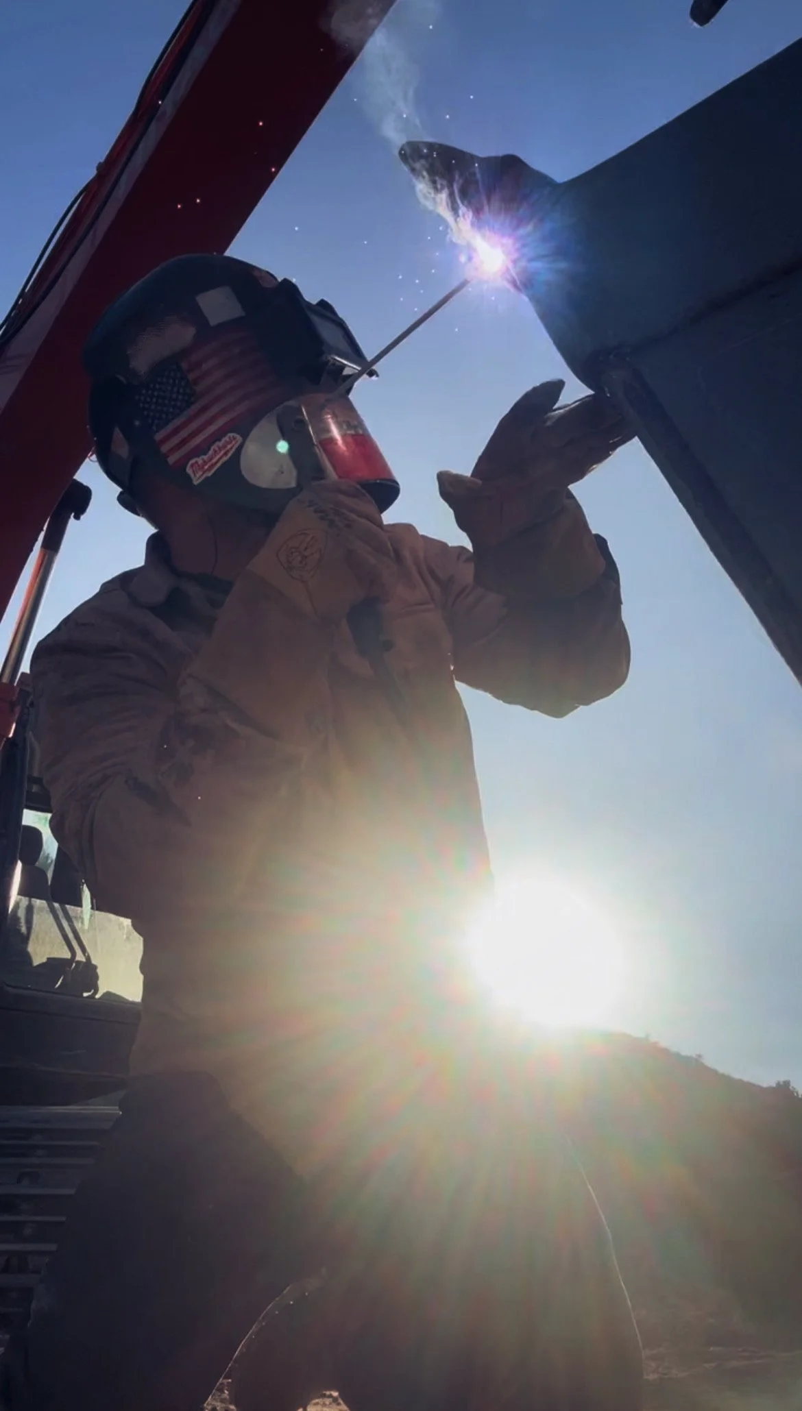 A person welding outdoors in bright sunlight, wearing protective gear including a helmet with an American flag sticker and gloves, welding a metal object.