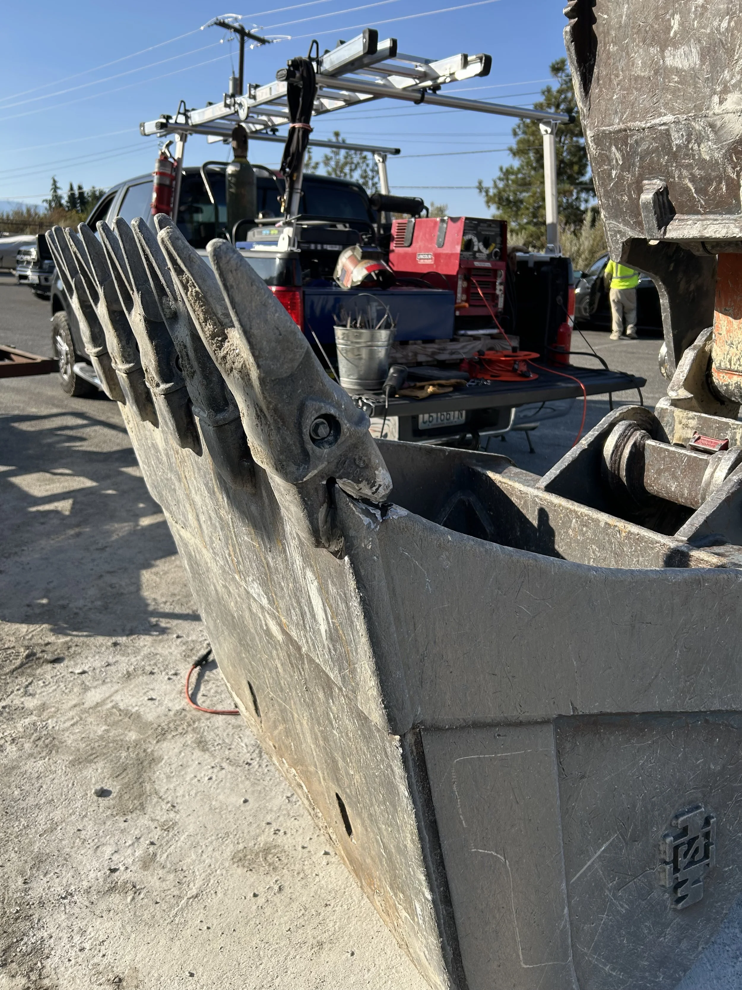Close-up of a large excavator bucket with dirt and wear, part of construction equipment, with a work site and vehicles in the background.