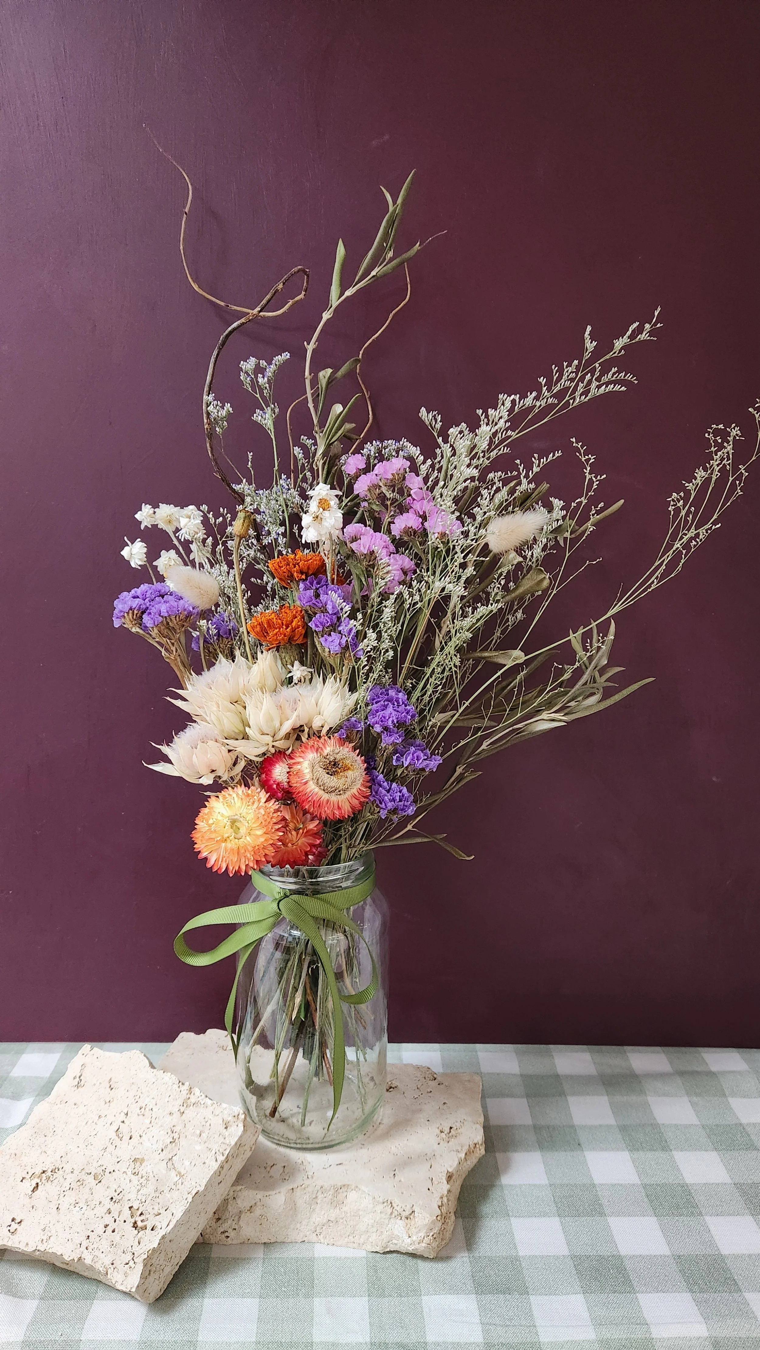 A glass vase with a bouquet of dried and fresh flowers, including purple, white, orange, and pink blooms, tied with a green ribbon, placed on two large, white stones on a green and white checkered tablecloth against a purple background.