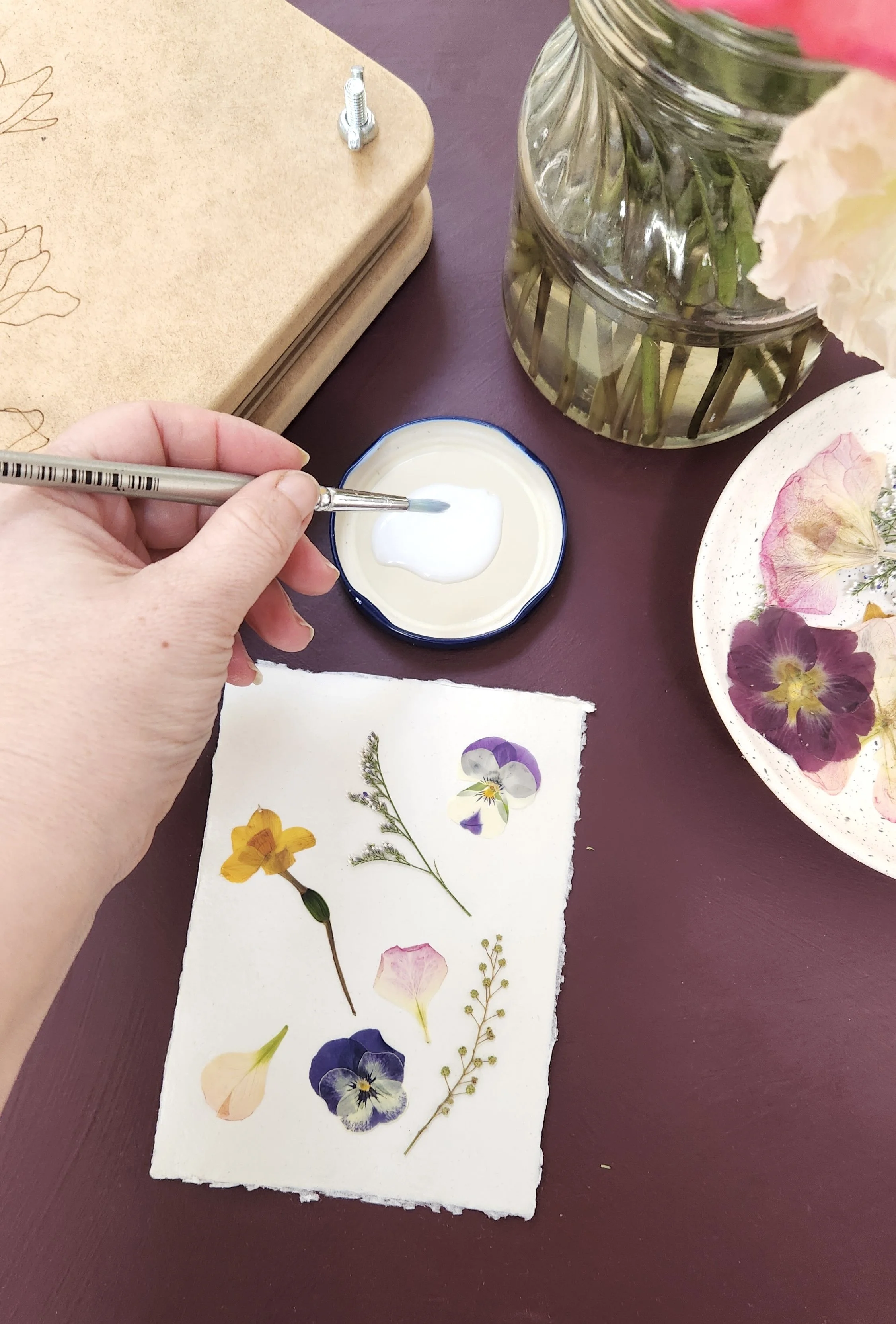 Person using a fine brush to apply a clear substance to pressed flower artwork on handmade paper, with a jar of water, dried flower petals, and a notebook on a maroon table.