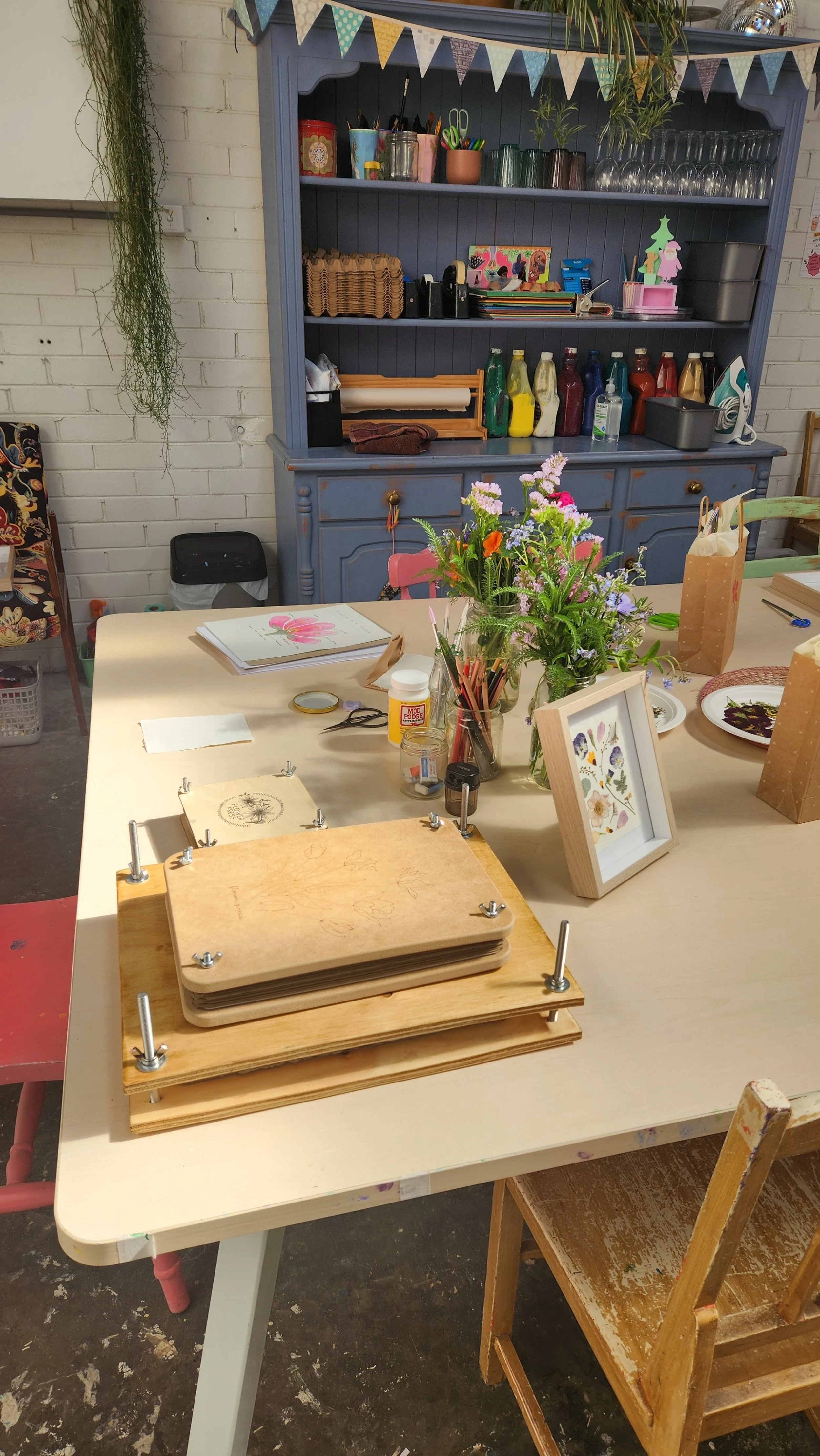 A decorated craft or art workspace with a table holding a flower arrangement, framed art, wooden boards, and art supplies. In the background, a blue shelf with cups, bottles, art supplies, and decorative items is against a white brick wall, with bunting hanging above.