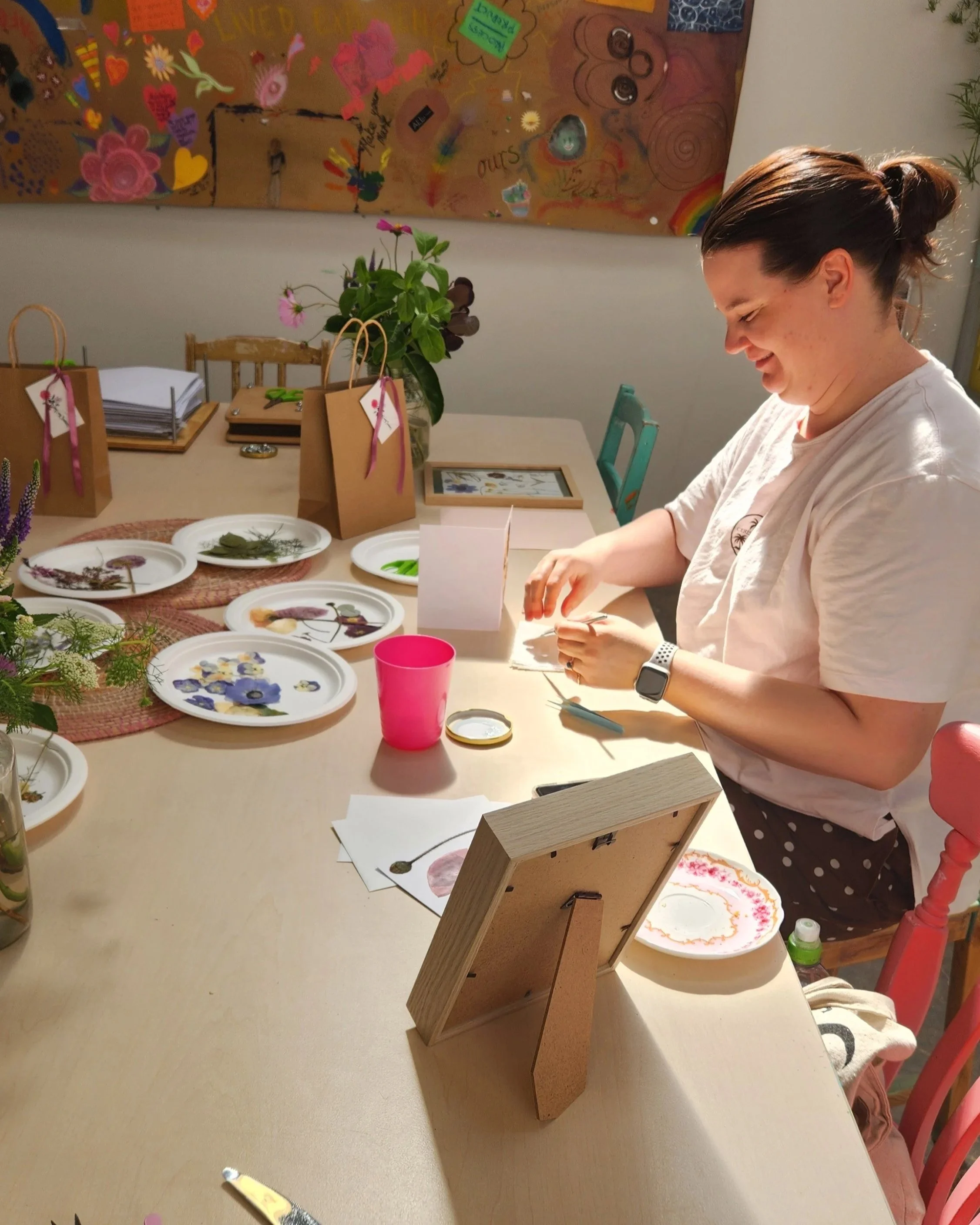 A woman sitting at a table with artwork, plates of pressed flowers, and crafting supplies, with a colorful decorated wall in the background.