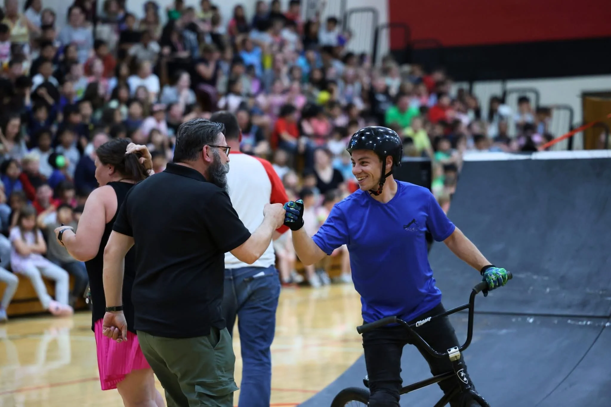 A young man smiling and wearing a helmet, riding a black scooter, shaking hands with a man and a woman in a gymnasium filled with spectators.