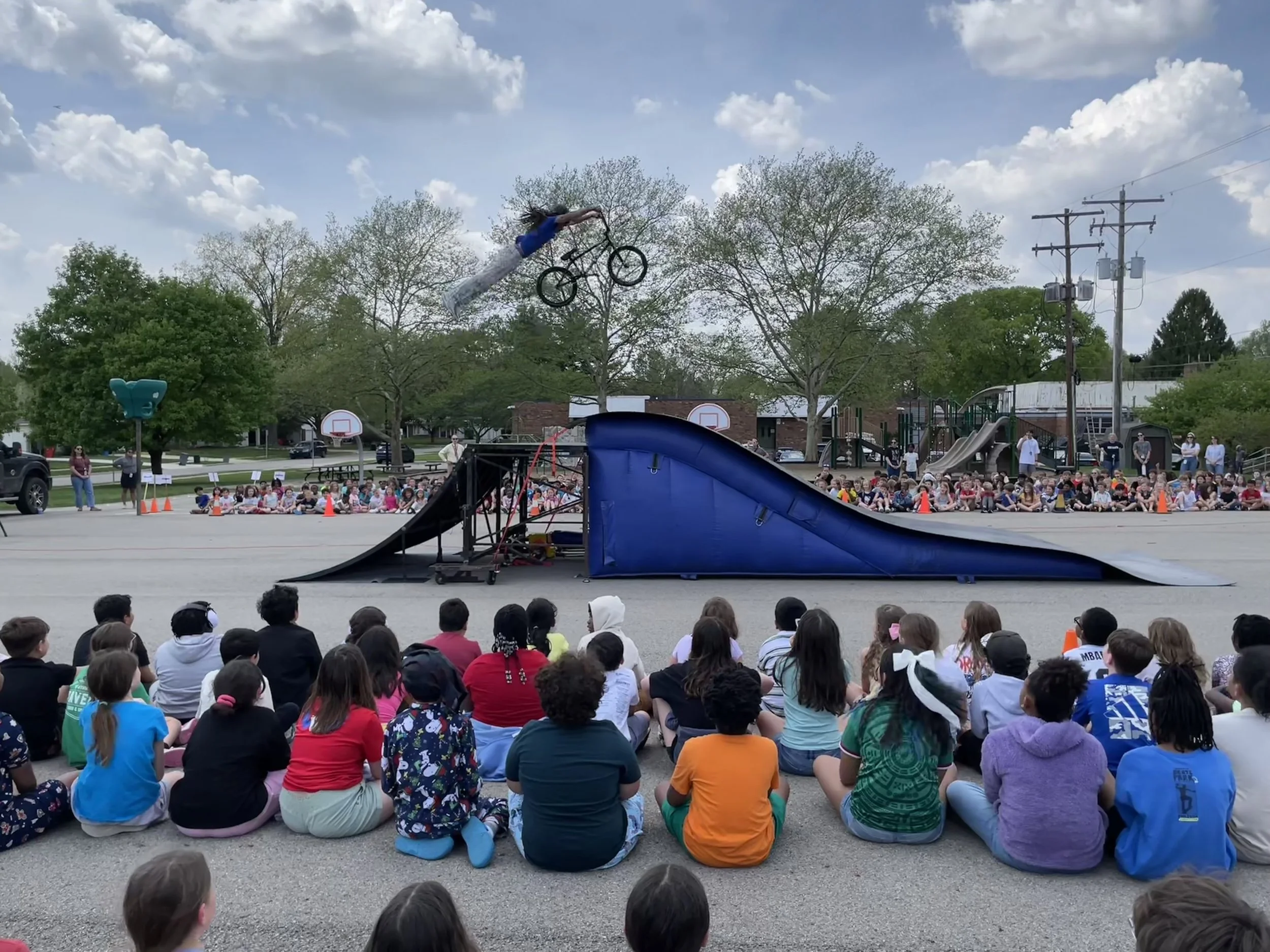 A young person is performing a bicycle stunt in the air at an outdoor event, with a large crowd of children and adults watching. The scene includes trees, utility poles, and playground equipment under a partly cloudy sky.