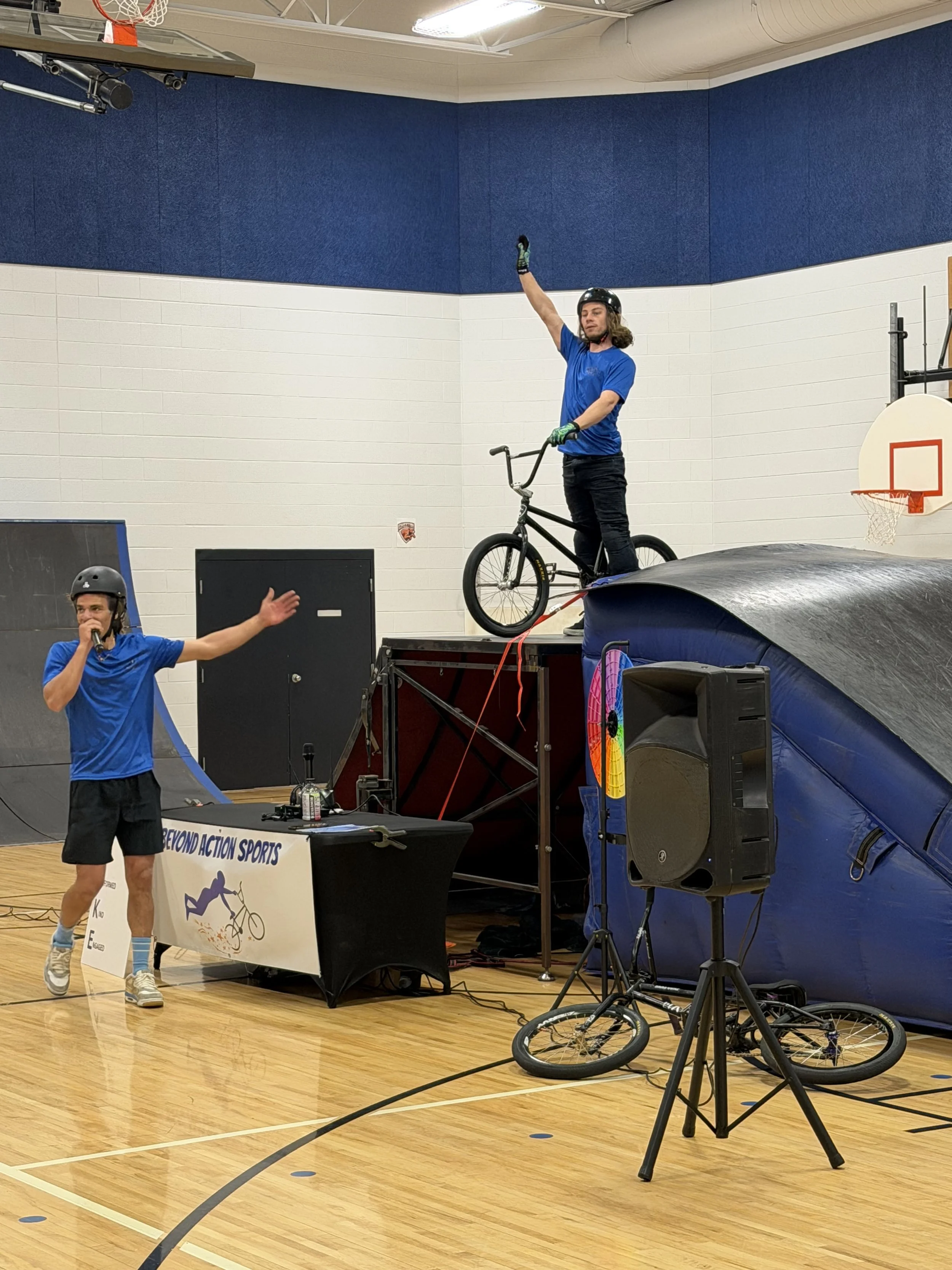 Indoor basketball gym with a person riding a BMX bike on a raised platform, another person speaking into a microphone, and various equipment including a trampoline, a bicycle, and a banner that reads 'BEYOND ACTION SPORTS'.