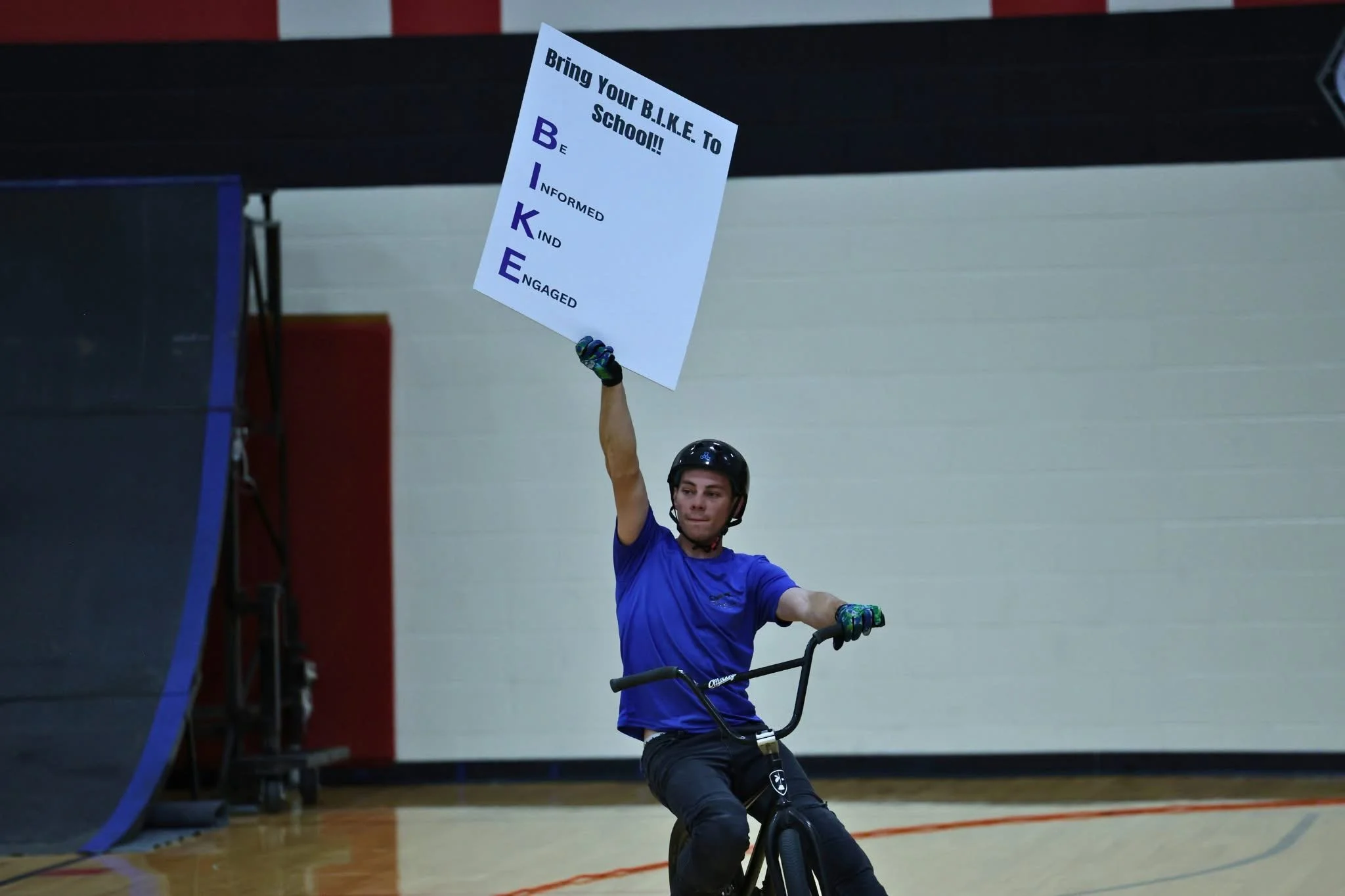 A young man in a helmet and blue shirt riding a unicycle inside a gymnasium. He is holding a sign with the word 'BIKE' spelled vertically, each letter representing a word: Be Informed, Kind, Engaged. The sign encourages bringing your bike to school.