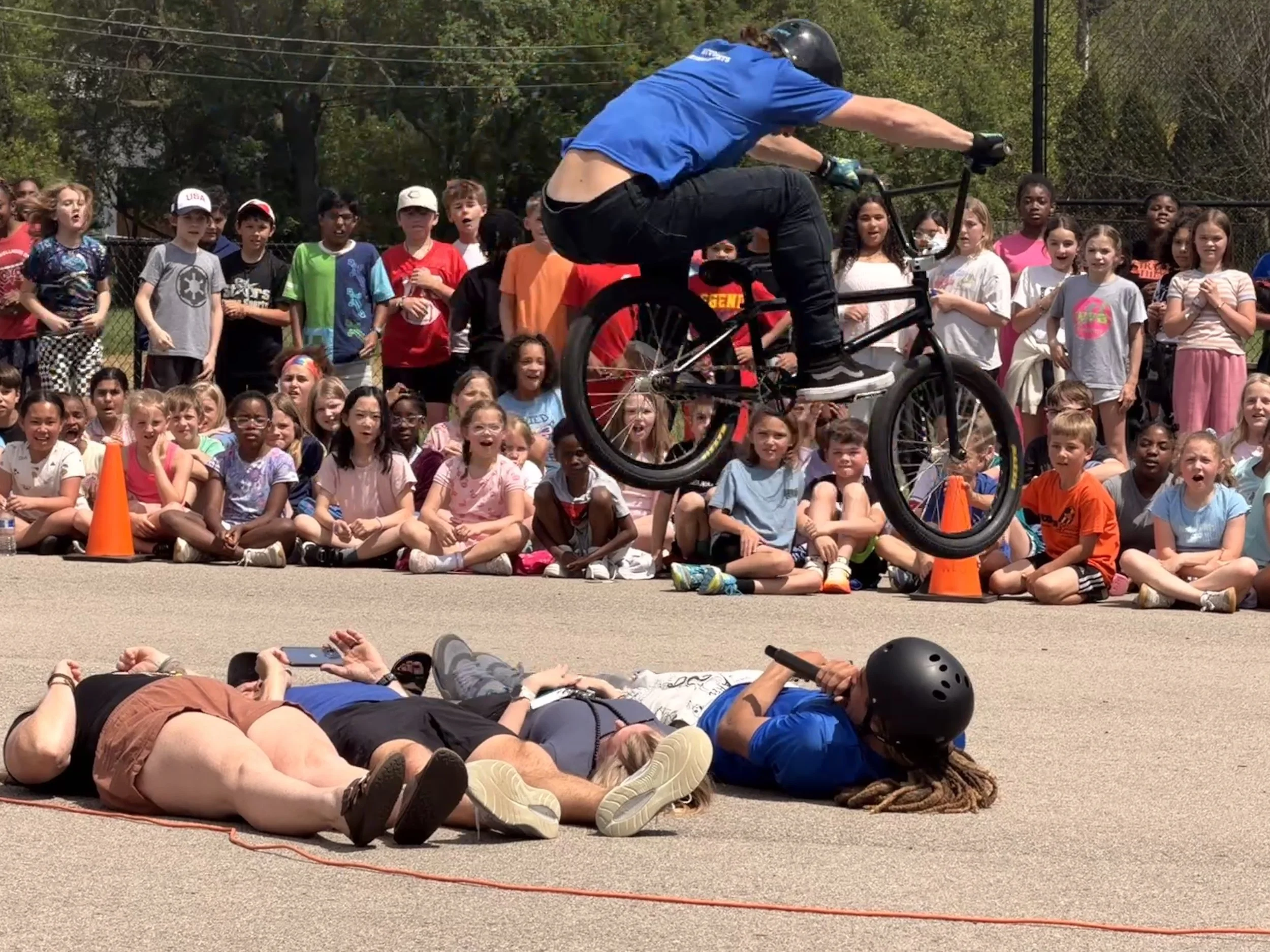 A crowd of children and adults watching a person riding a bicycle on a sunny day at a park or outdoor event.