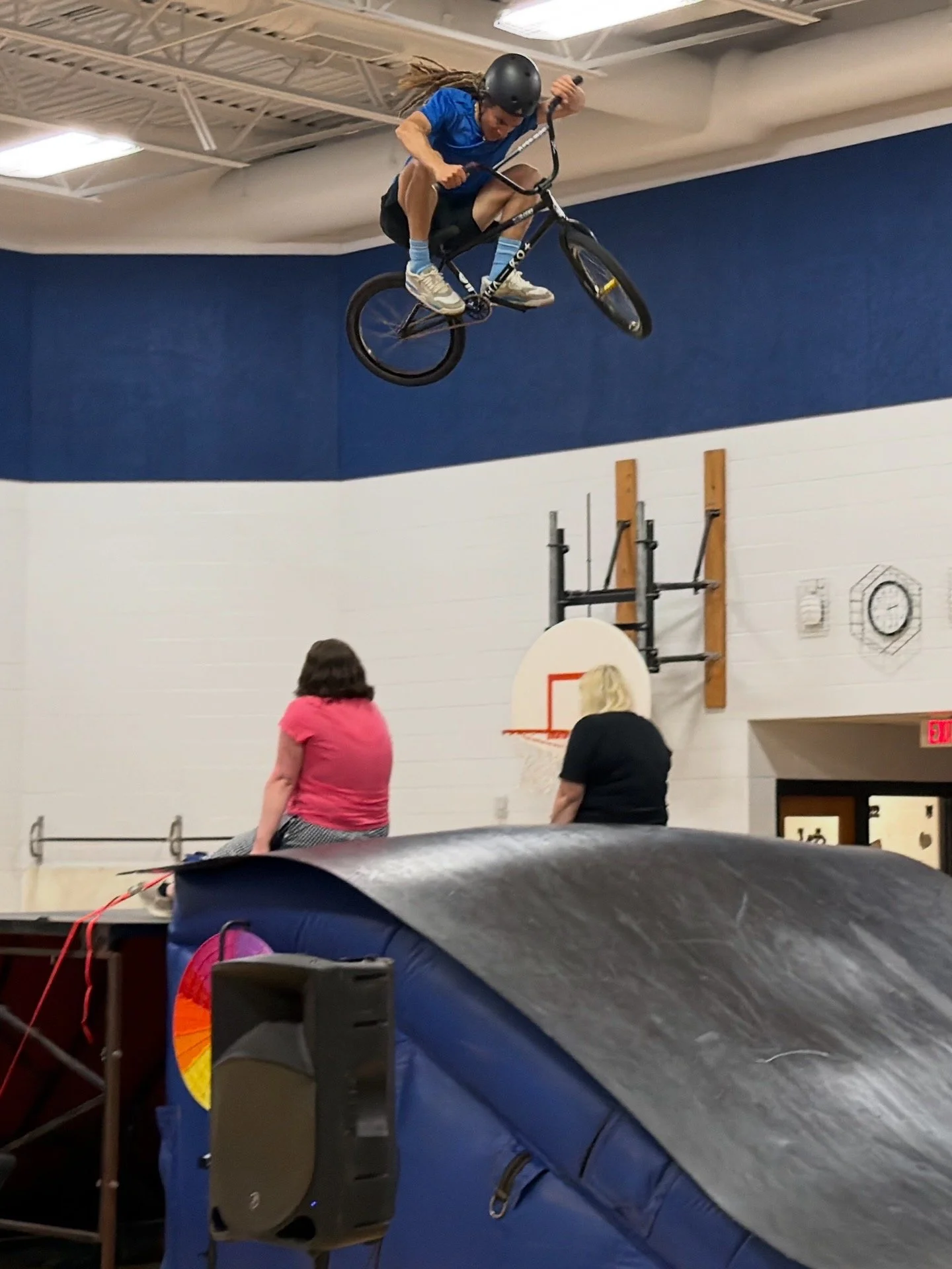 Person performing a BMX trick in an indoor gym, mid-air above a padded area, with spectators watching.