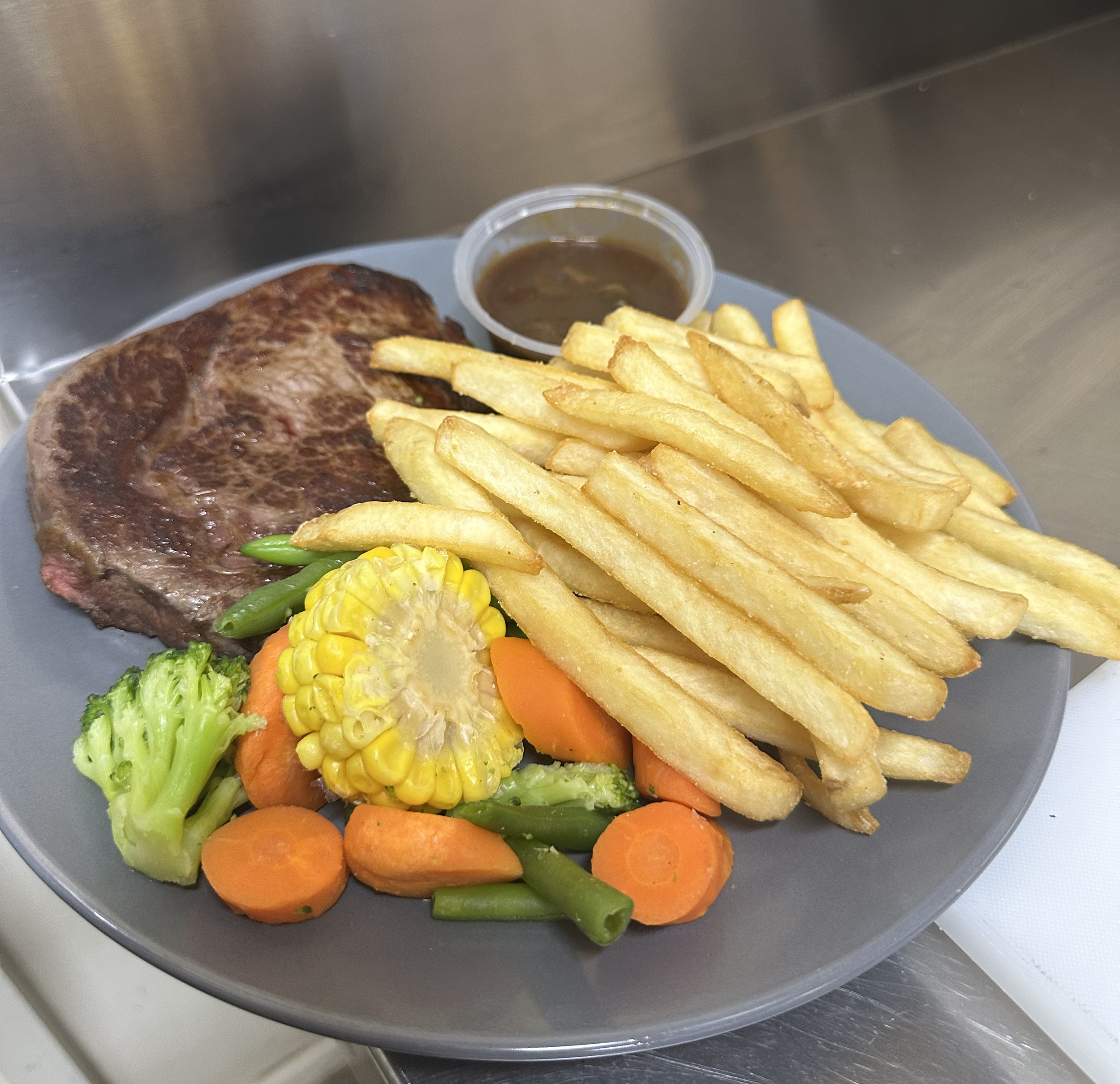 Plate of cooked steak, French fries, mixed vegetables including broccoli, carrots, corn, and green beans, with a small cup of brown gravy.