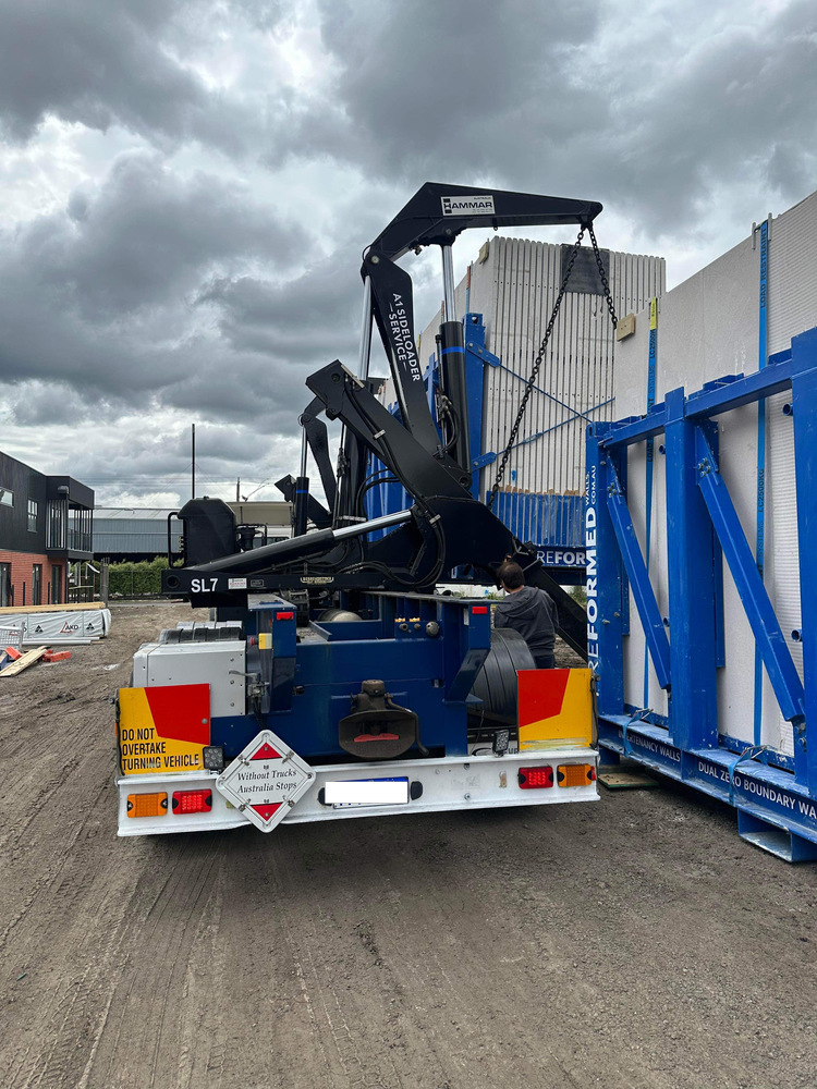 Truck with a side loader crane lifting construction materials outdoors on a cloudy day.
