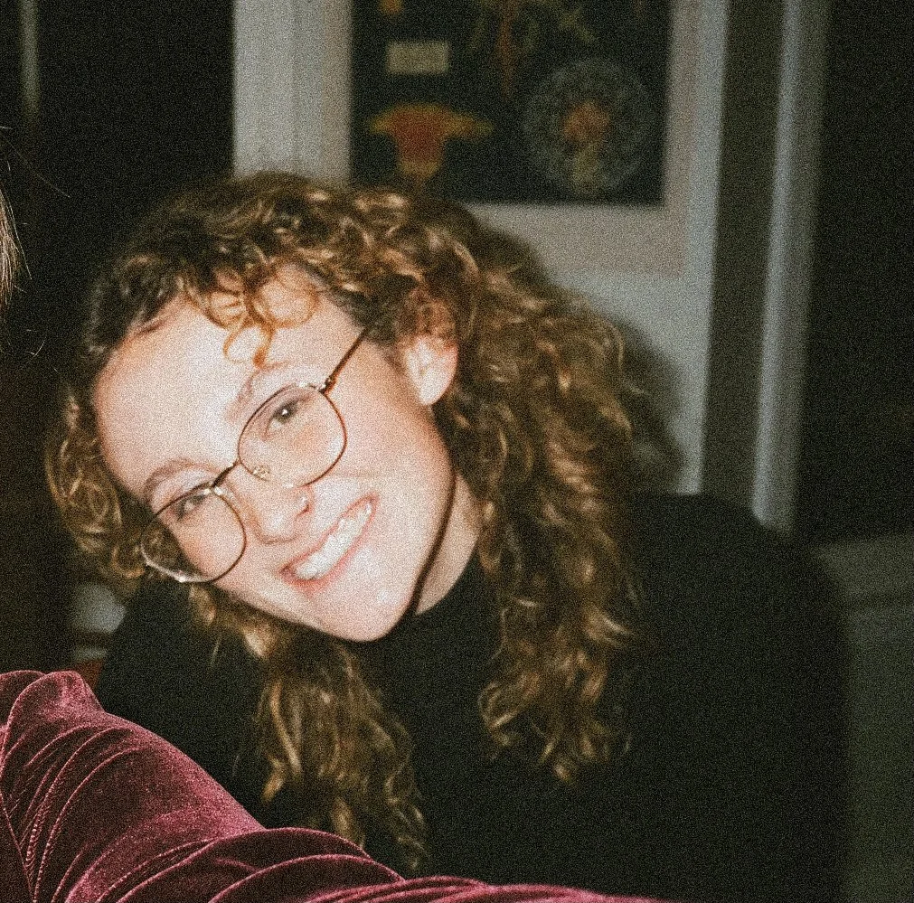 Person with curly hair and glasses smiling indoors, wearing a black top.