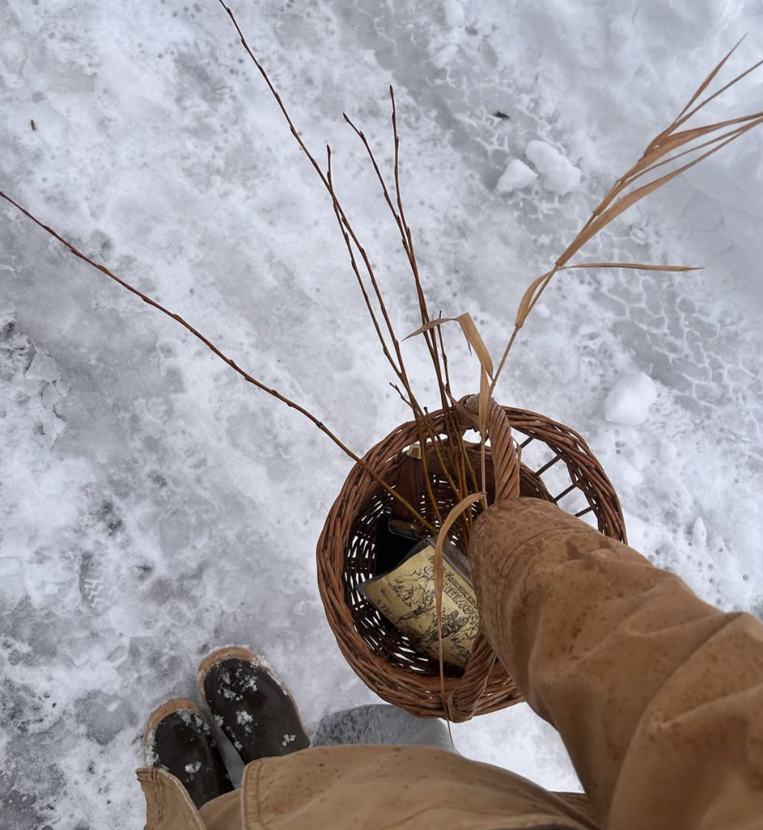 Person wearing brown boots and coat standing on snowy ground, holding a wicker basket with branches and a decorated bottle.