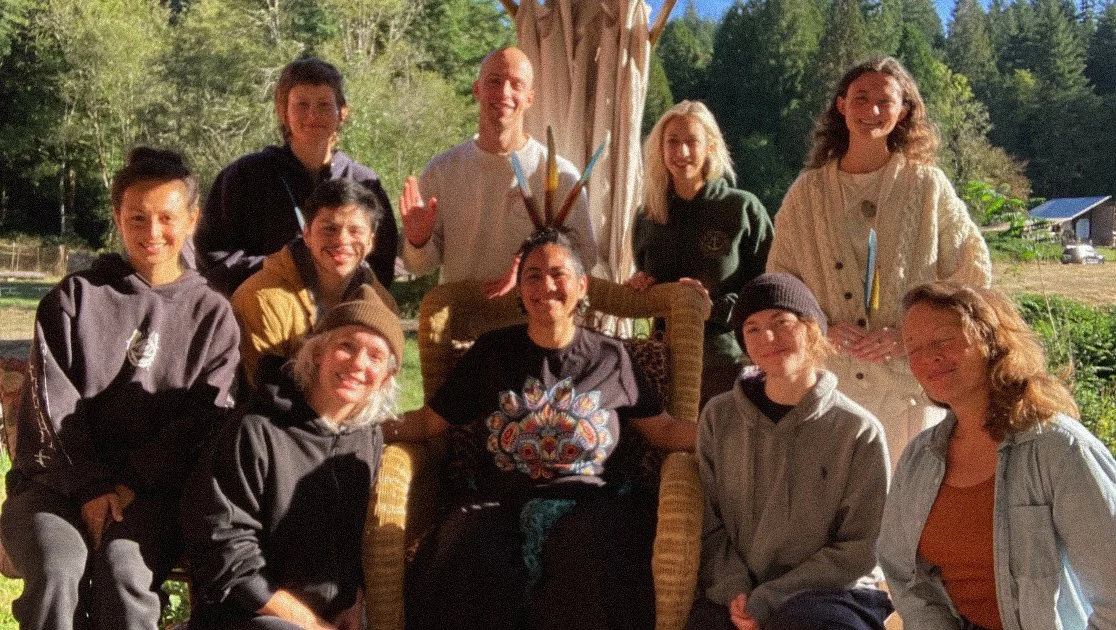 A group of people posing outdoors, smiling and gathered around a person seated on a woven chair. They appear to be in a natural setting with trees and grass in the background. The group is casually dressed, and the person in the chair has colorful decorations resembling feathers behind them.