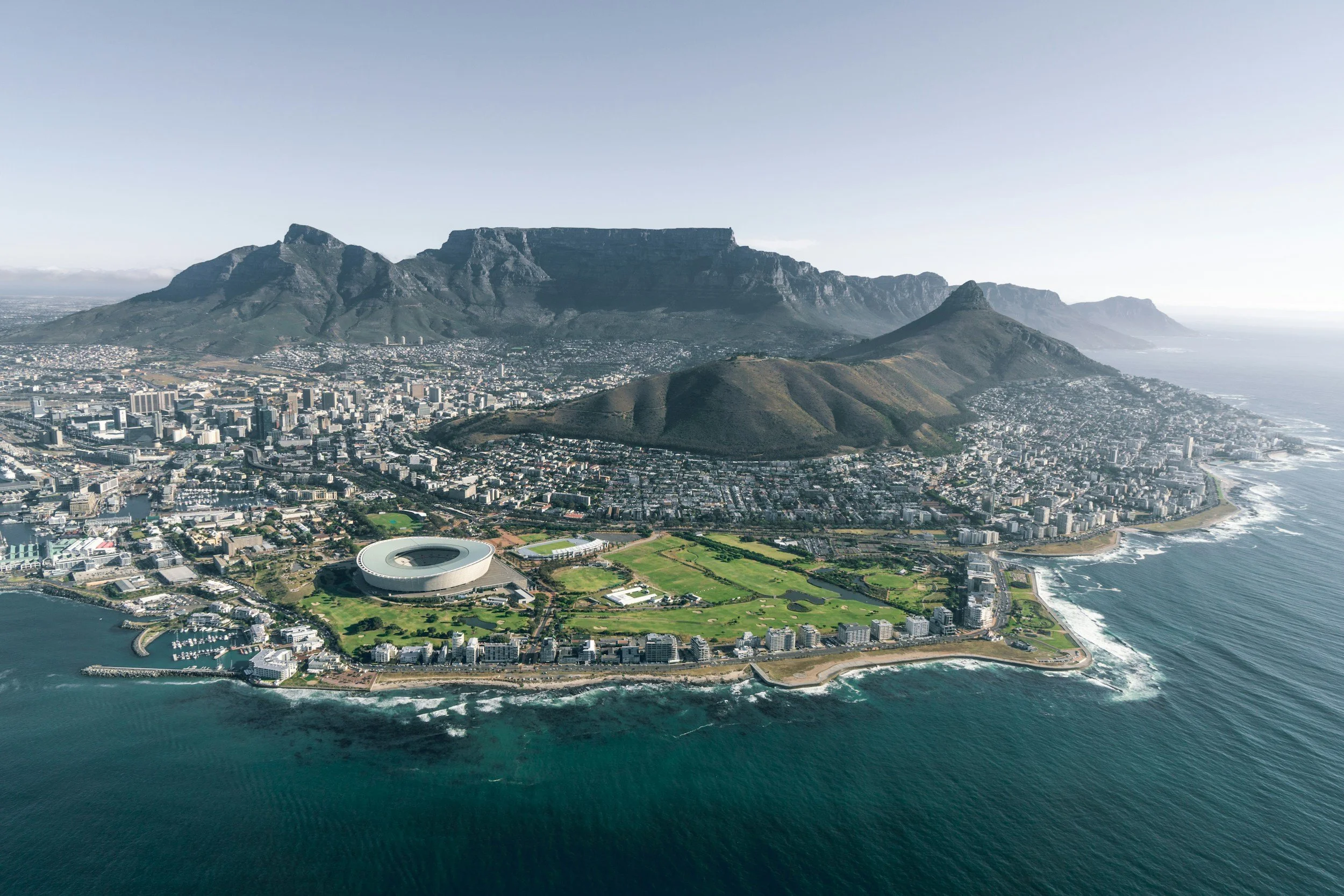 An aerial view of Cape Town, South Africa, featuring the cityscape, Table Mountain, and Robben Island in the background, with the Atlantic Ocean in the foreground.