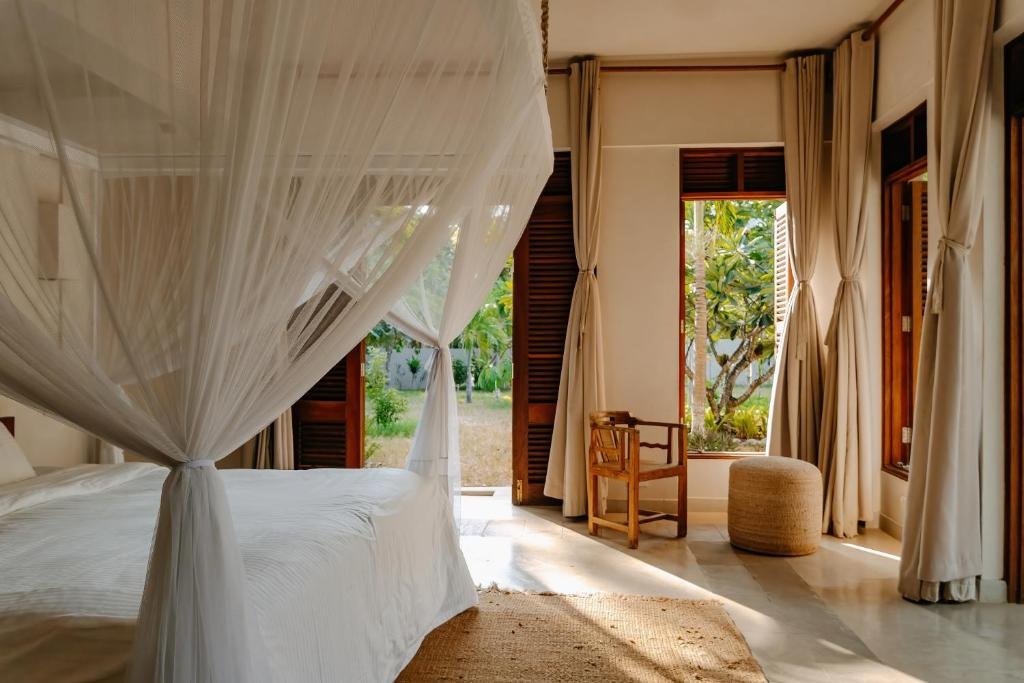 A bedroom with a large bed with a canopy and white curtains, overlooking a lush outdoor garden through open windows.