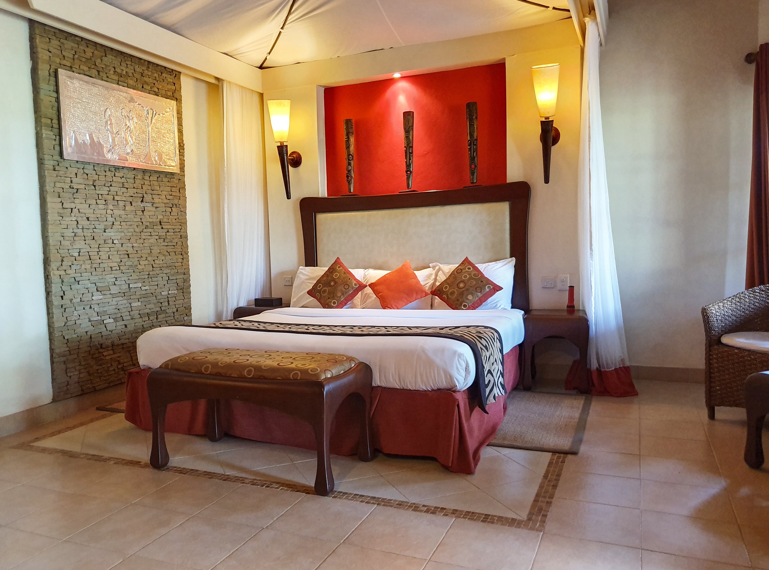 A hotel room featuring a large bed with decorative pillows, a bench at the foot of the bed, and a bedside table. The room has a mix of modern and rustic decor, with wall sconces, a textured stone wall, and curtains on the windows.