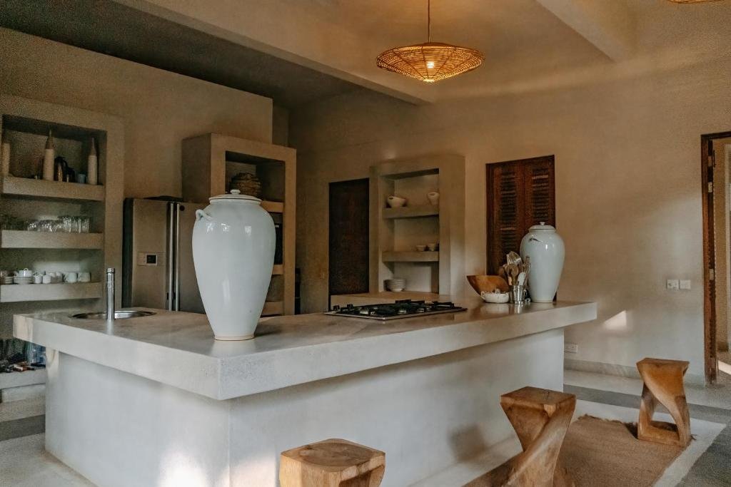 Modern kitchen with white counters, two large white vases, wooden stools, open shelving, small silver refrigerator, beige walls, and a woven ceiling light.
