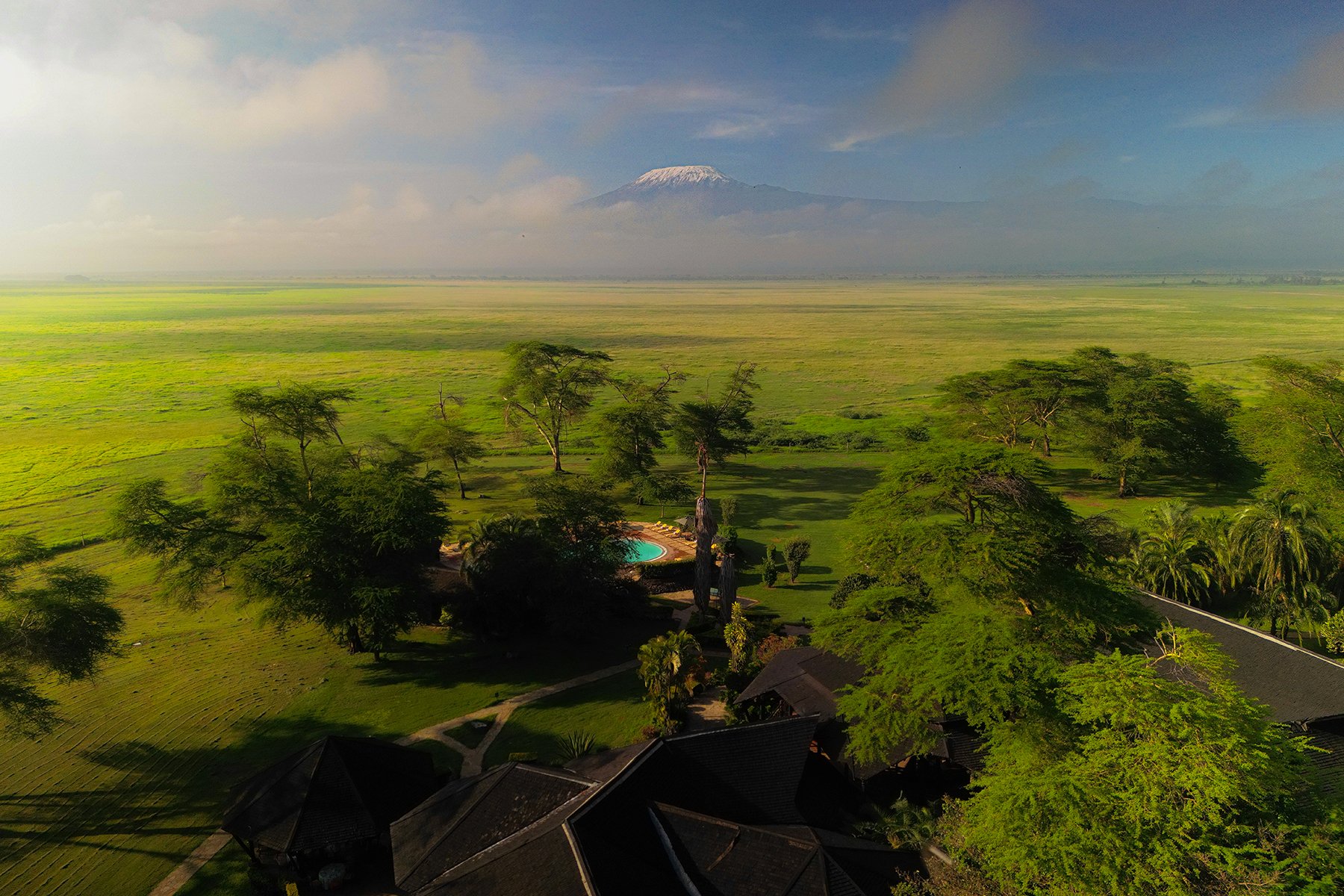 Aerial view of lush green landscape with tall trees, a small pool, and a building with dark rooftops in the foreground, expansive grassy fields in the middle ground, and a mountain with a snow-capped peak in the background.