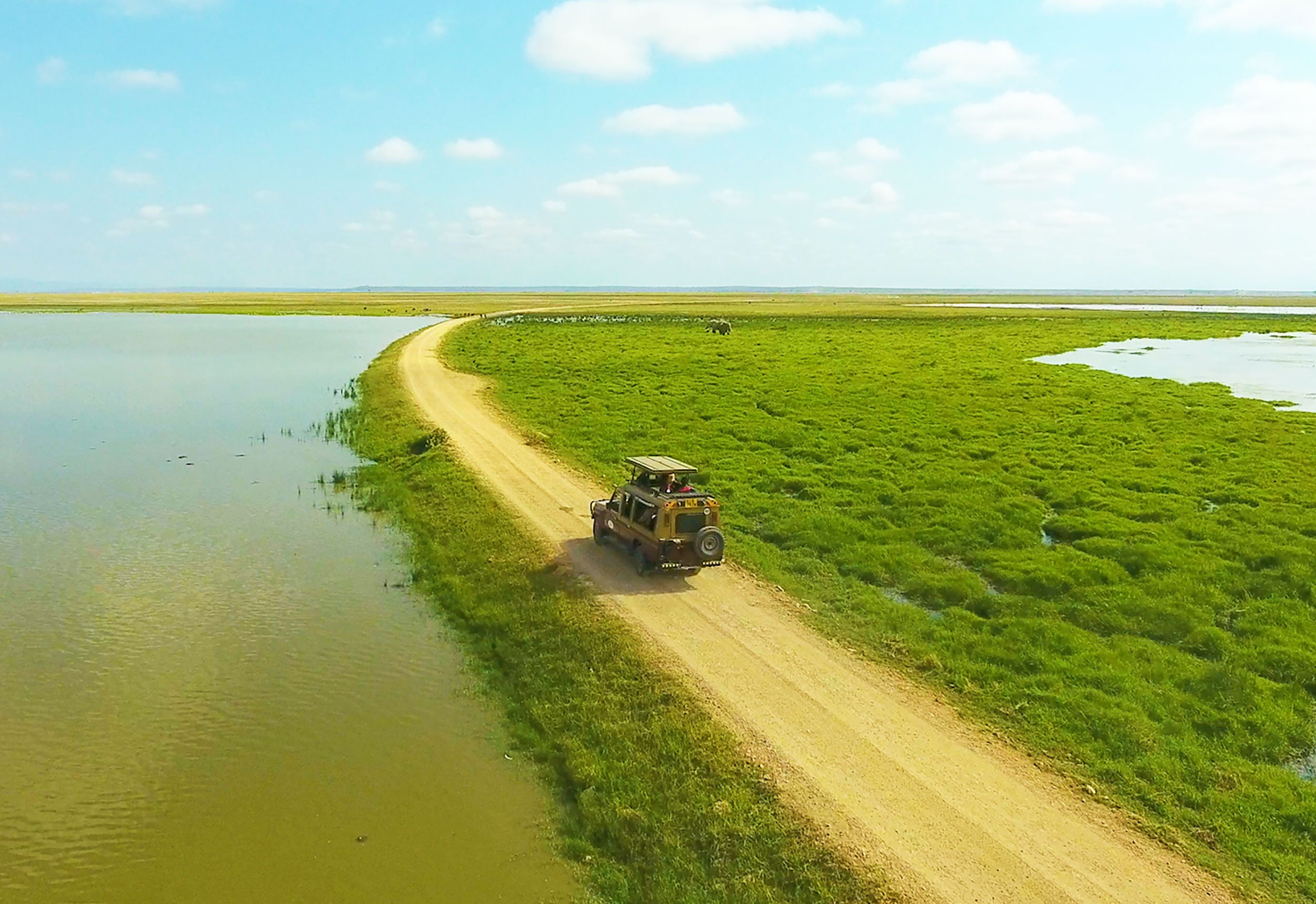 An off-road vehicle driving on a dirt road between a body of water on the left and green marshland on the right, under a partly cloudy sky.