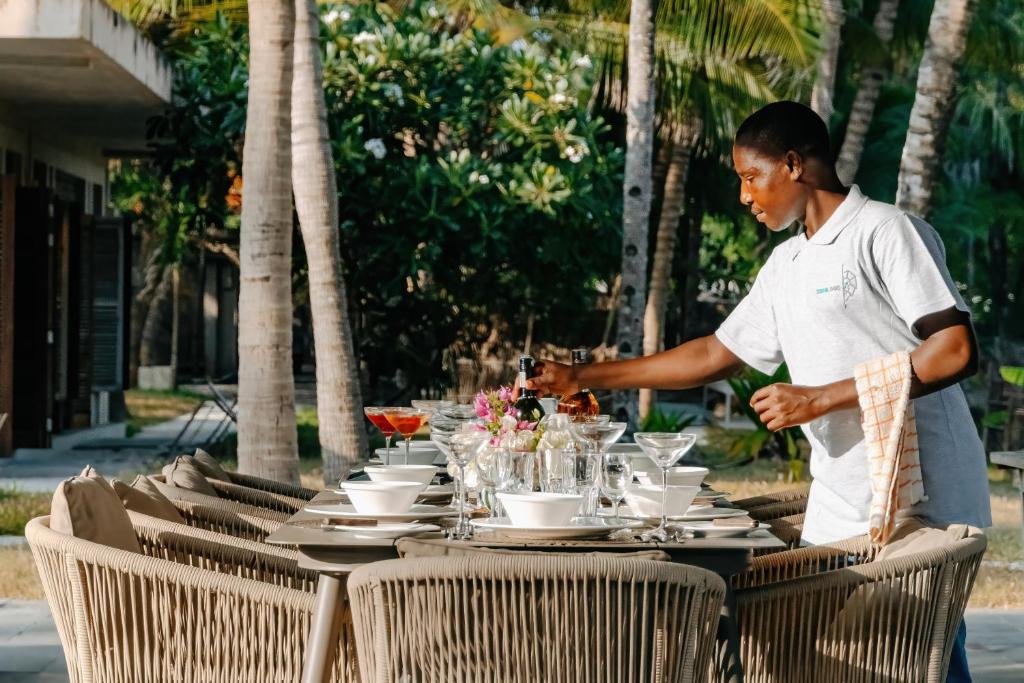 A person setting an outdoor dining table with glassware and flowers in a tropical setting with trees and a building in the background.