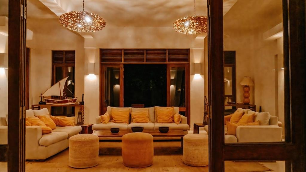 Living room with beige sofas, yellow pillows, two round ottomans, wooden furniture, a ship model on a side table, and warm lighting.