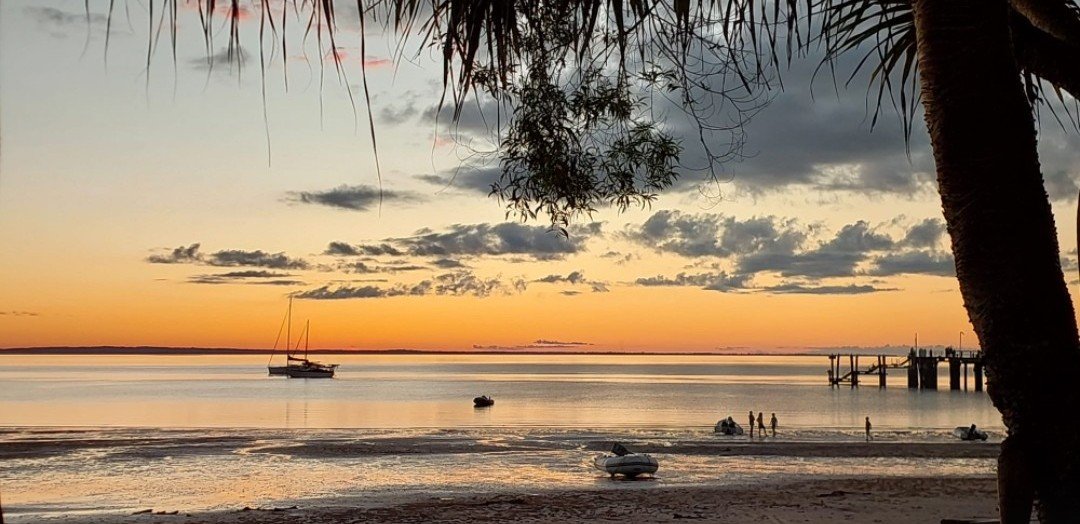 Outdoor dining table with eight wicker chairs around it, set with placemats, glasses, and a fruit bowl in a wooded area.
