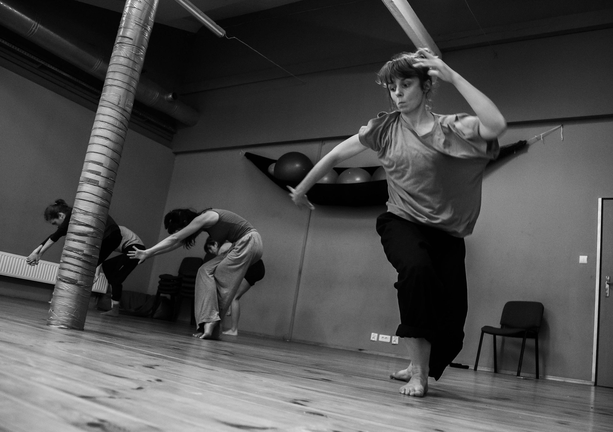 A participant balances with an outstretched arm while others explore movement in a wooden-floor studio.