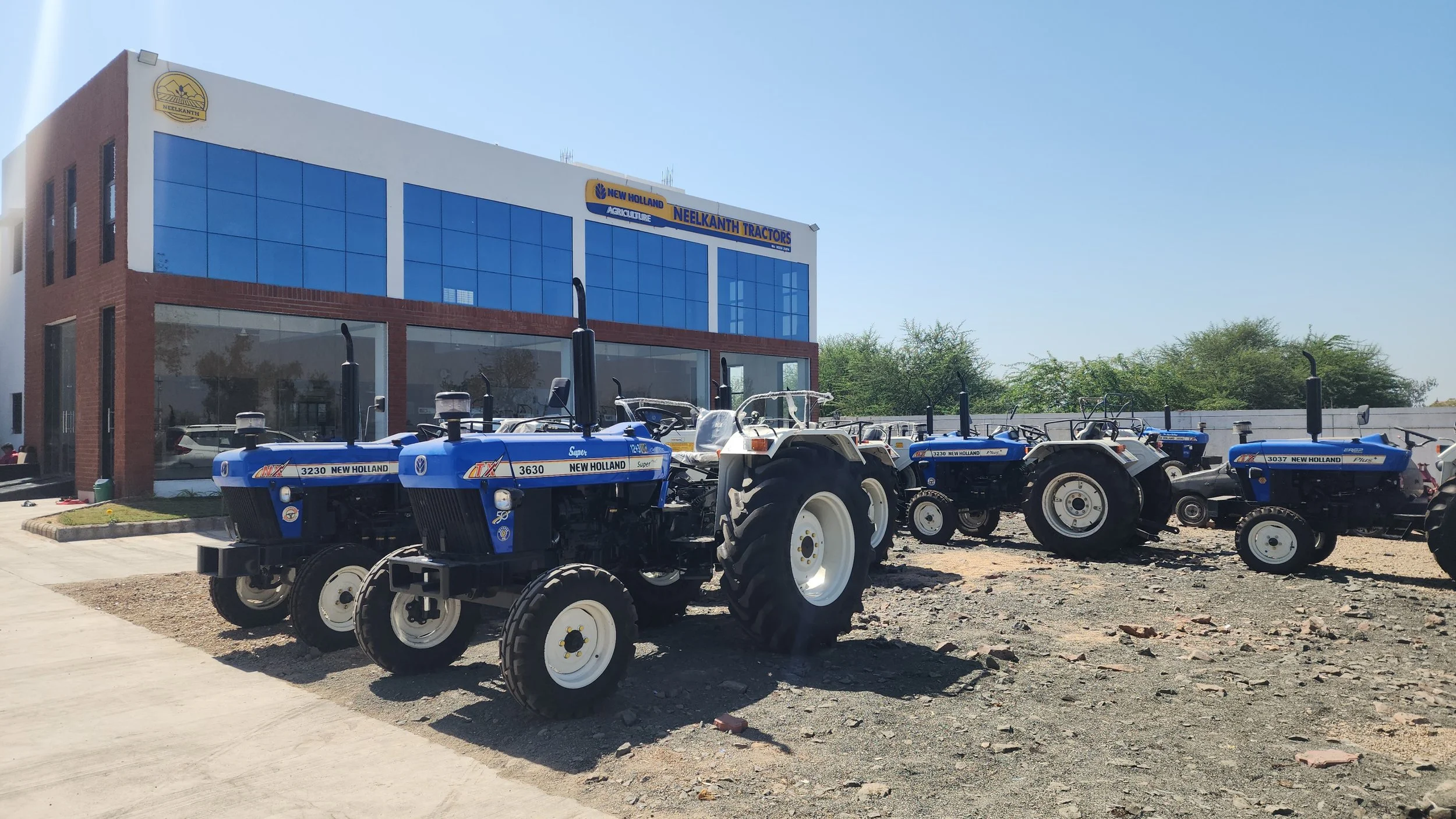 A row of New Holland Tractors parked outside a modern building with reflective glass windows and signage for Neelkanth Tractors and New Holland Tractors. The scene is set on a clear, sunny day with some trees in the background.
