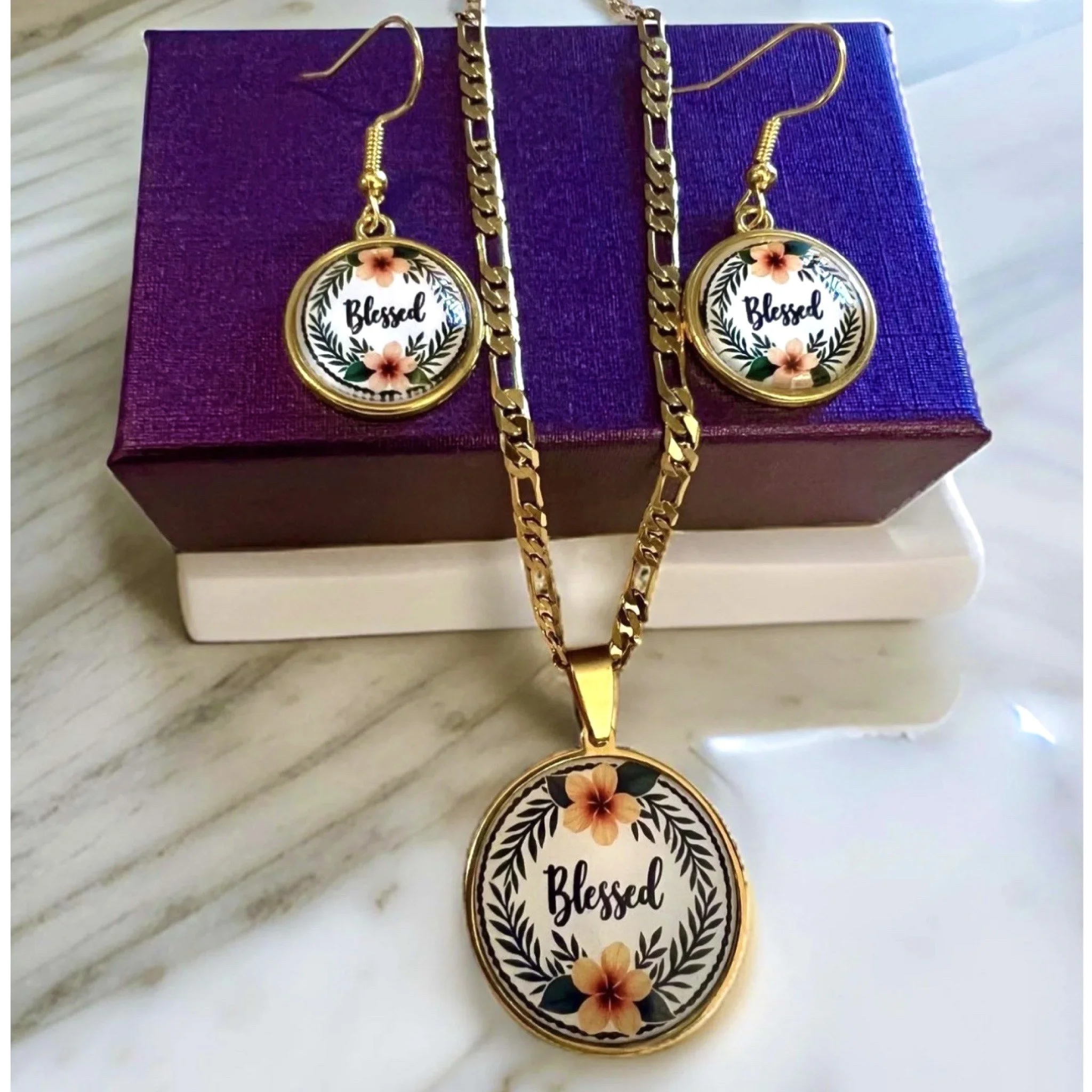 Gold jewelry set including a necklace and earrings with 'Blessed' inscription, floral and leaf design, displayed on a purple box and white background.