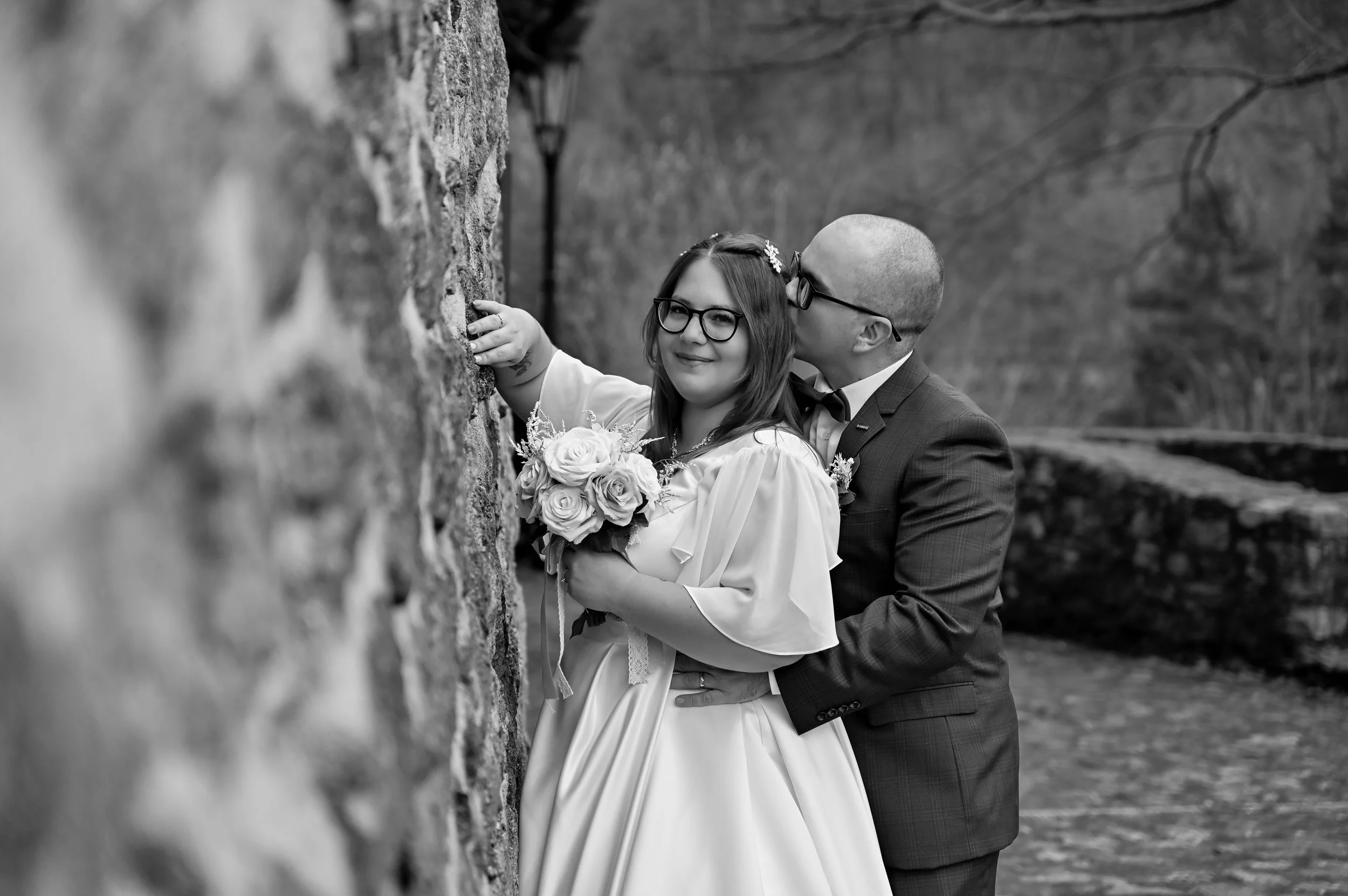 A couple dressed in wedding attire, the bride holding a bouquet of roses and the groom gently kissing her cheek, standing outdoors next to a stone wall.
