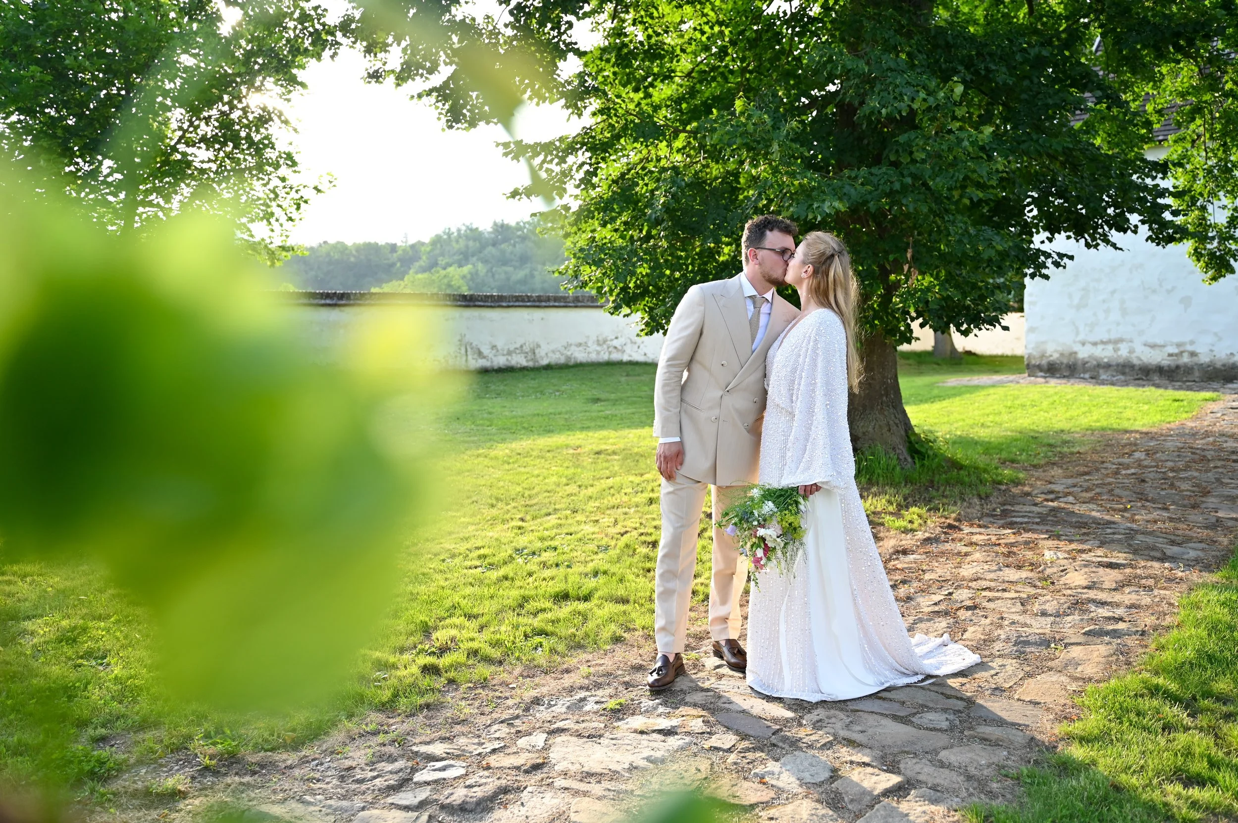 A couple dressed in wedding attire sharing a kiss outdoors near a large tree, with a white wall and green landscape in the background.