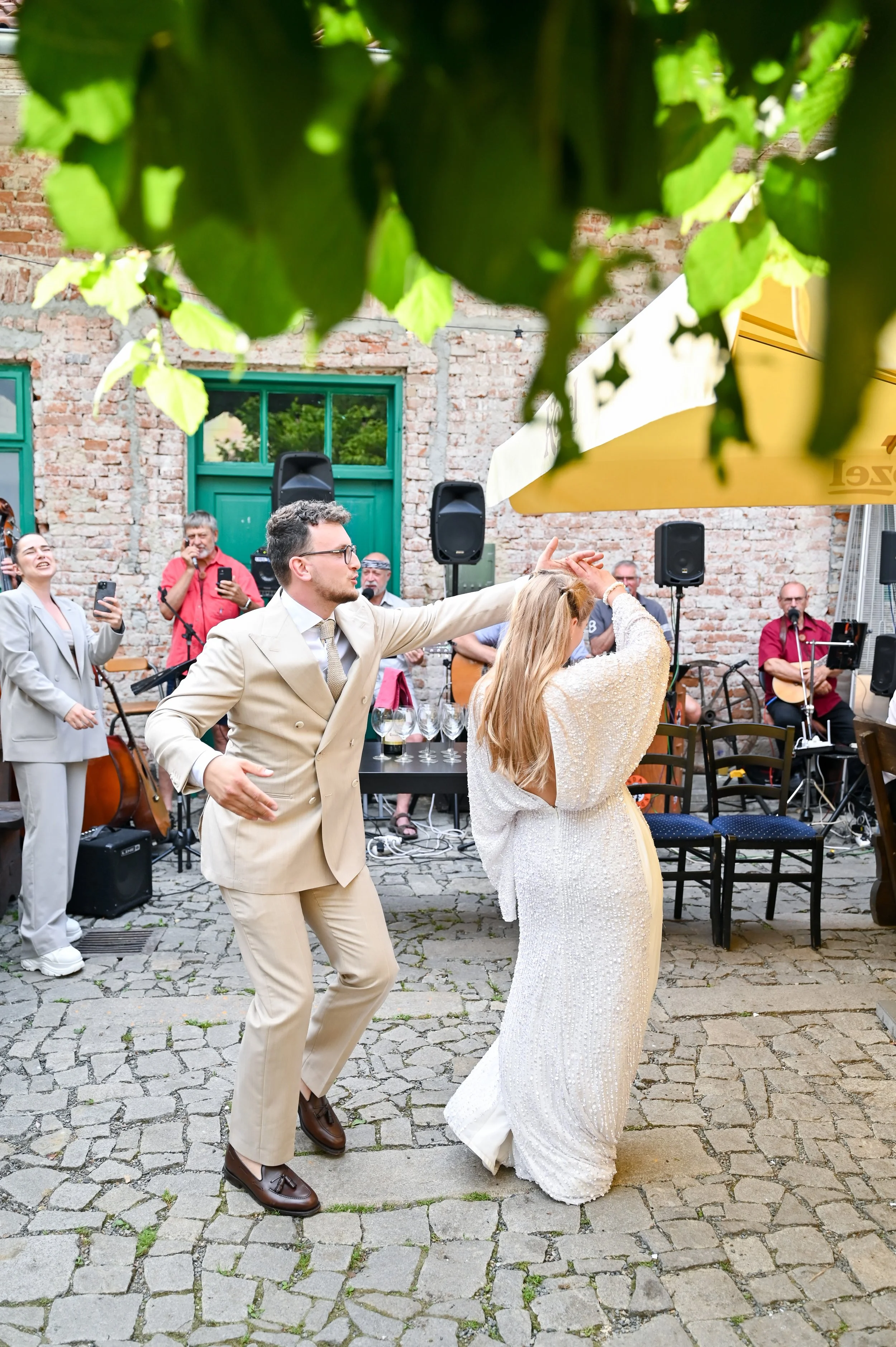 Couple dancing at an outdoor wedding reception, with a live band playing in the background, cobblestone ground, and green leaves partially visible at the top.