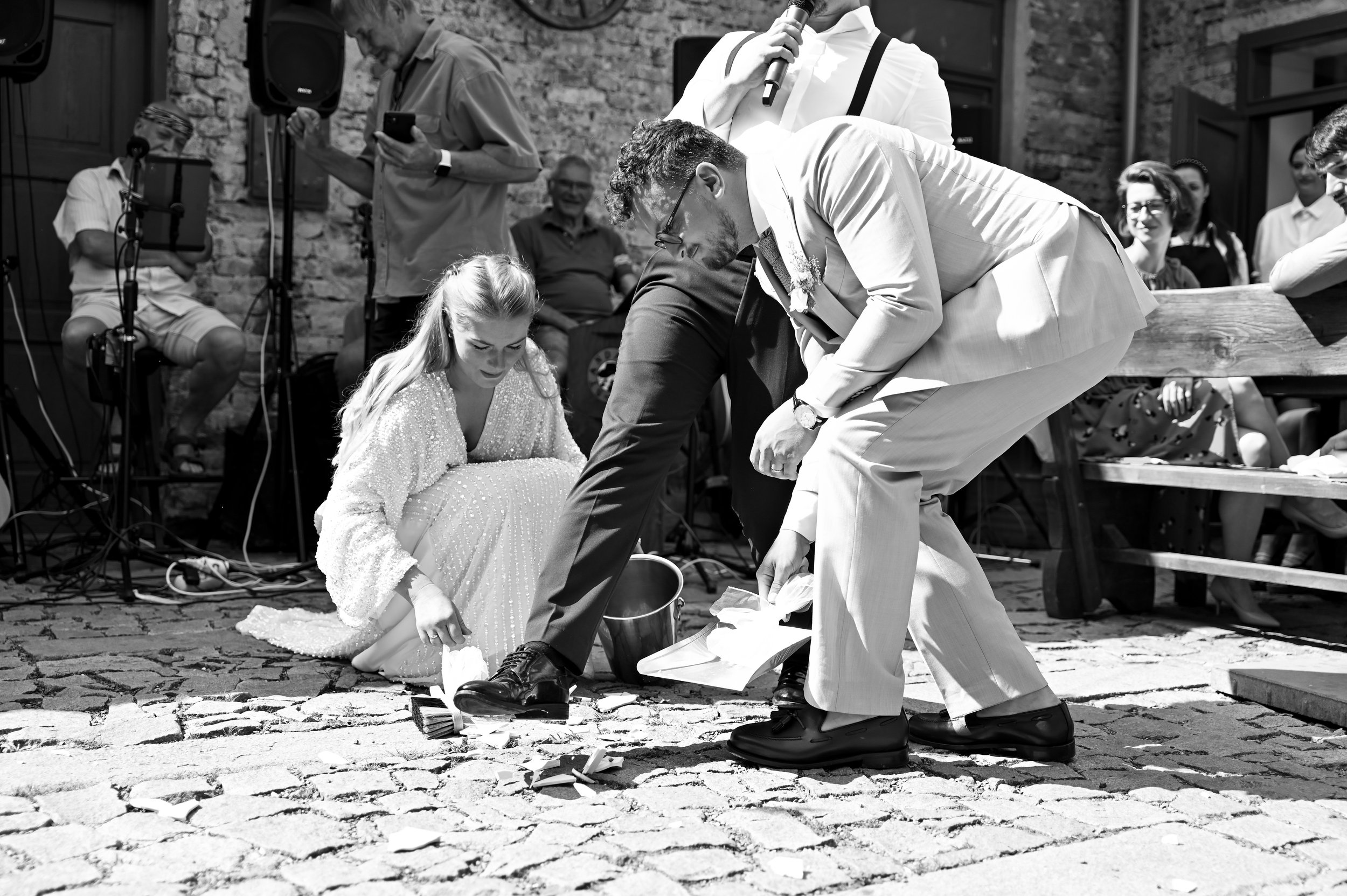 A black-and-white photo of a wedding ceremony where a couple is participating in a sand or flower ceremony, with several guests watching. The bride is kneeling on the ground in a sequined dress, and the groom is bent over, both pouring items from a c