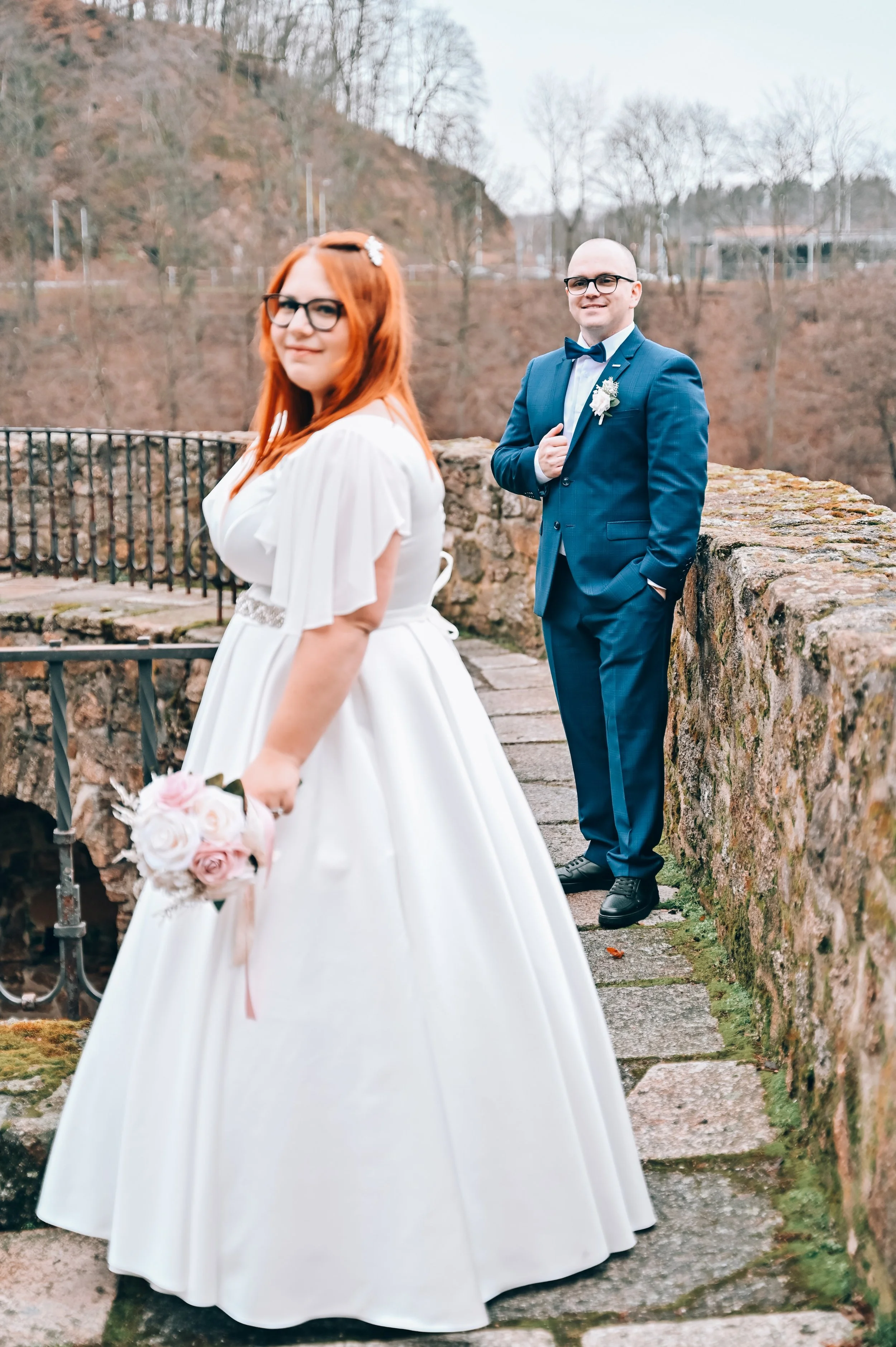 A bride in a white gown holding a bouquet of pink and white roses, and a groom in a blue suit with a bow tie, standing outdoors on a stone pathway with a stone wall and bridge in the background.
