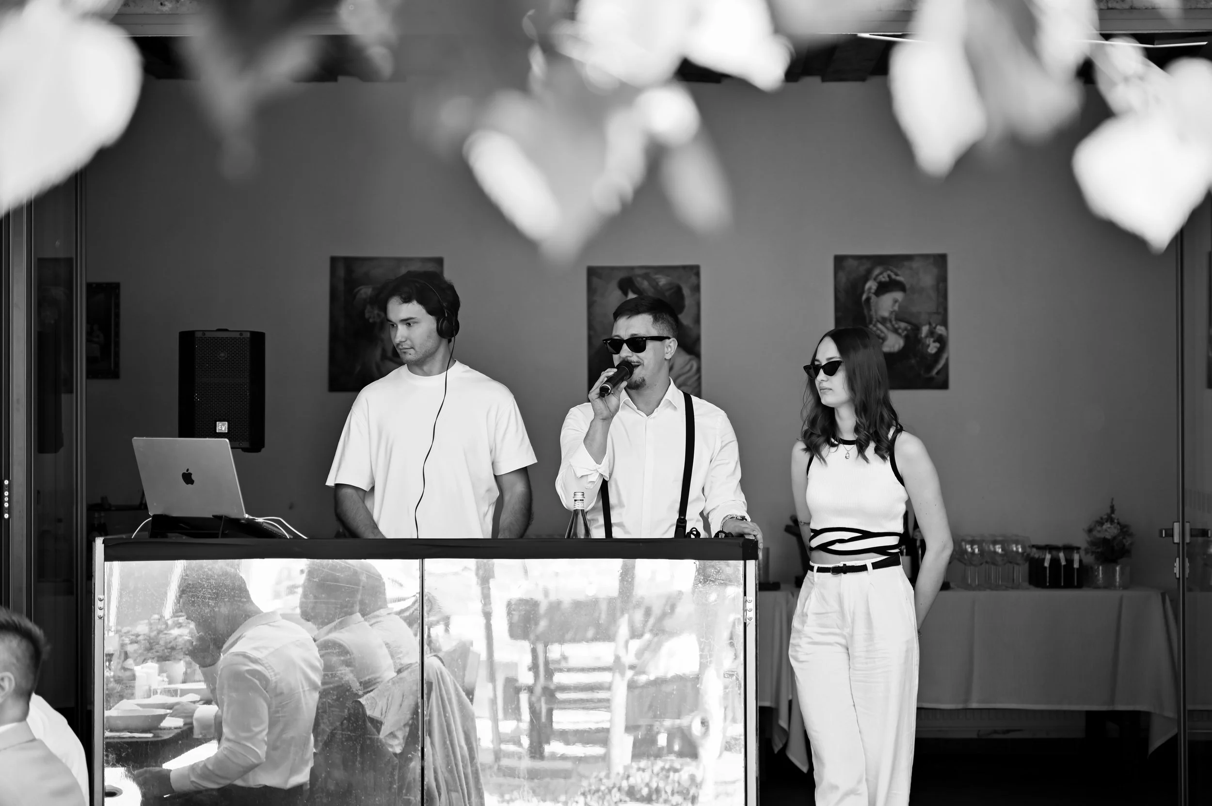 Three people during a performance at an event. The person in the middle is singing into a microphone, wearing sunglasses, a white shirt with suspenders. The person to the left is wearing headphones, working on a laptop. The person to the right is sta