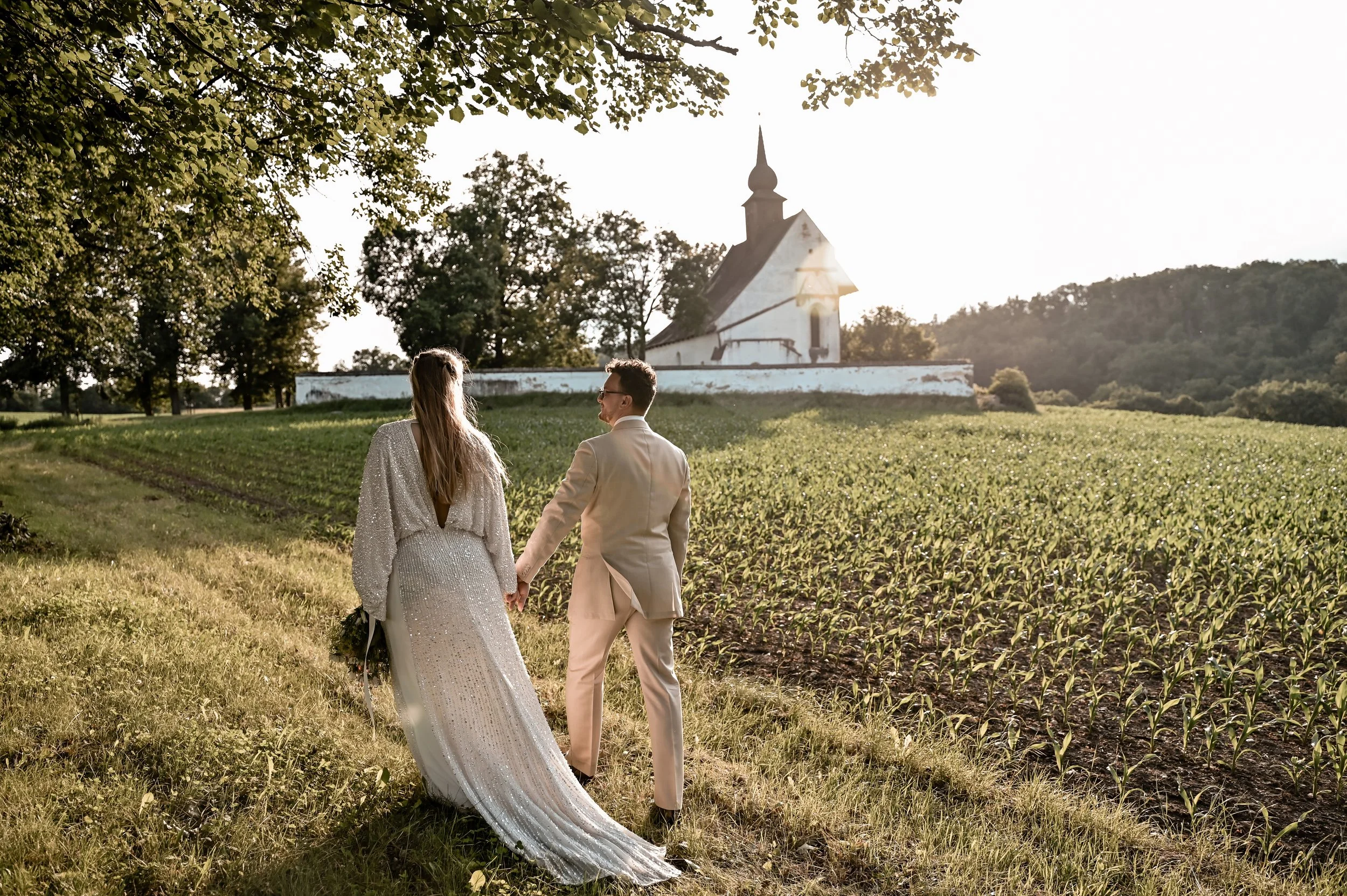 A bride and groom holding hands and walking through a grassy field towards a white church on a sunny day.