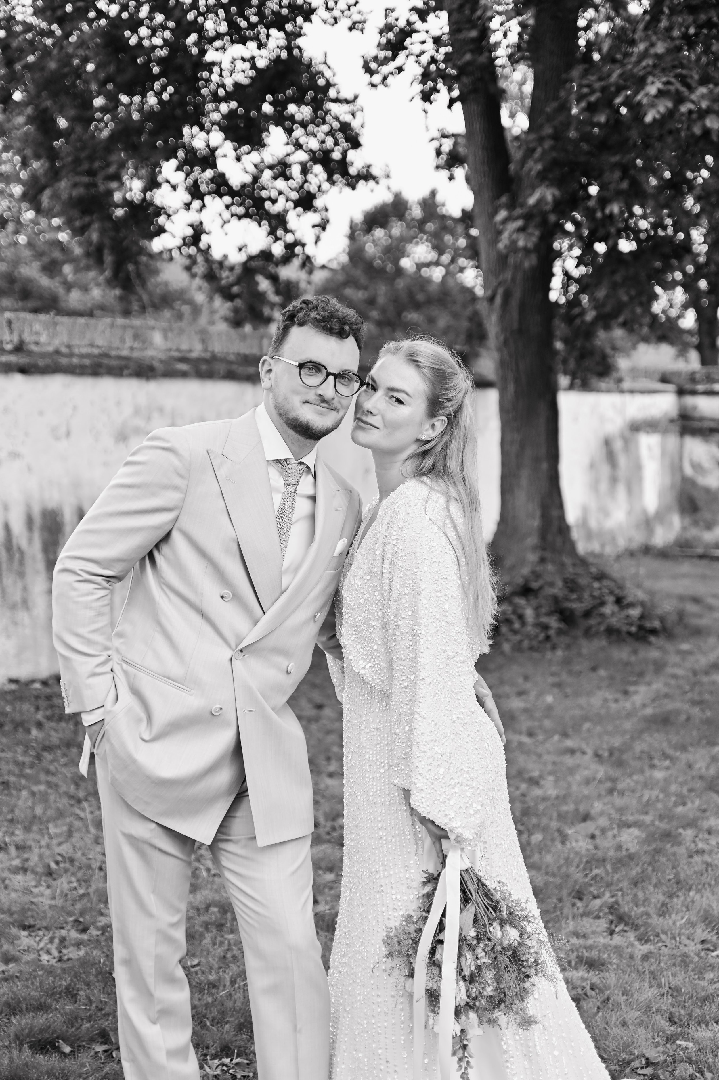 Black and white photo of a bride and groom standing outdoors near a tree and a stone wall, smiling and posing together on their wedding day.