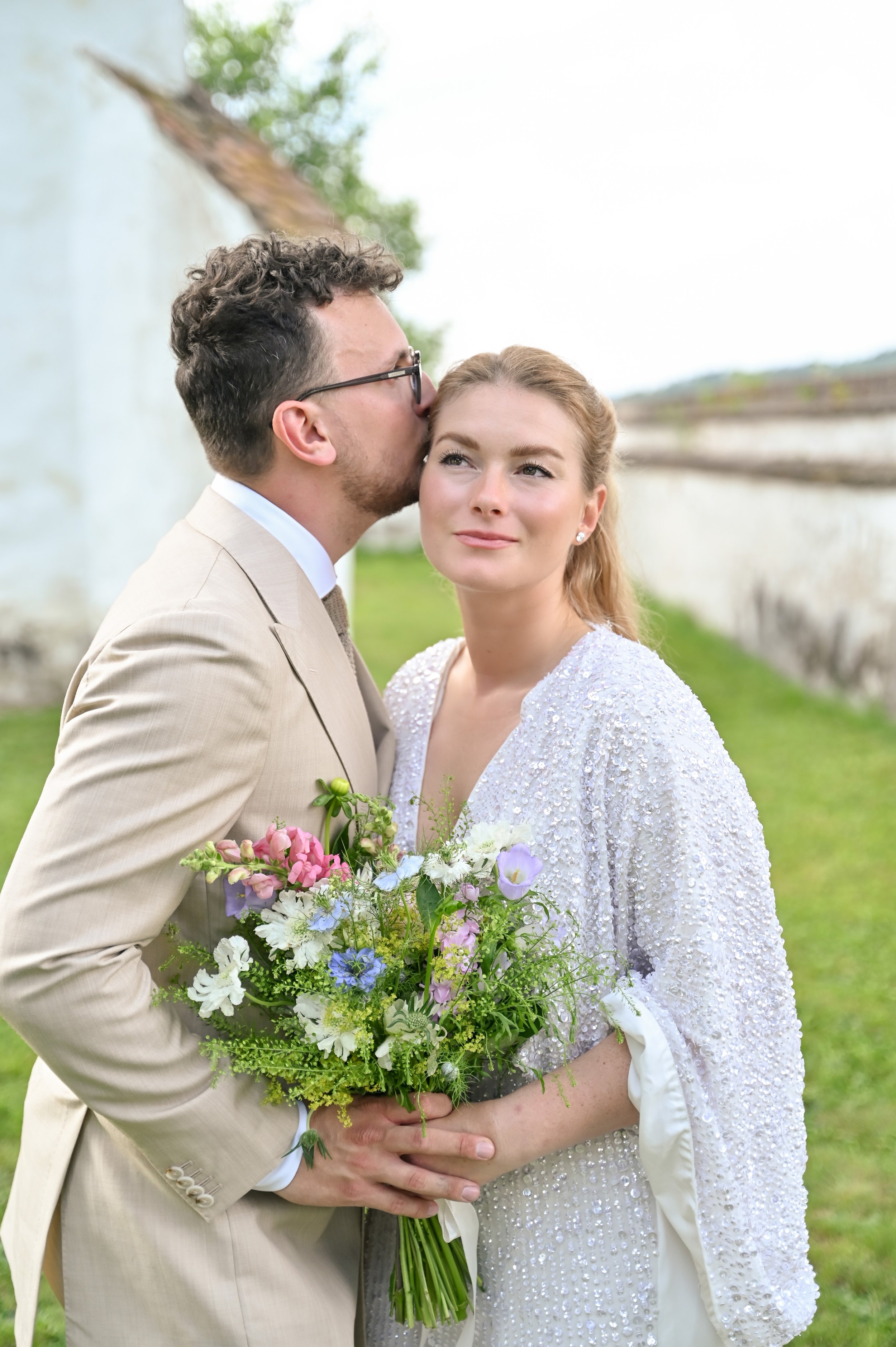 A bride holding a bouquet of flowers standing outside while a groom kisses her on the temple.