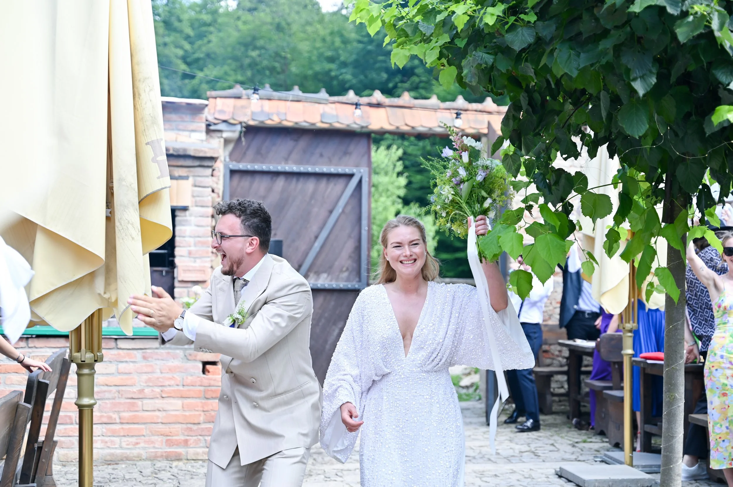 A bride holding a bouquet and a man in a suit clapping at an outdoor wedding reception with guests in the background.