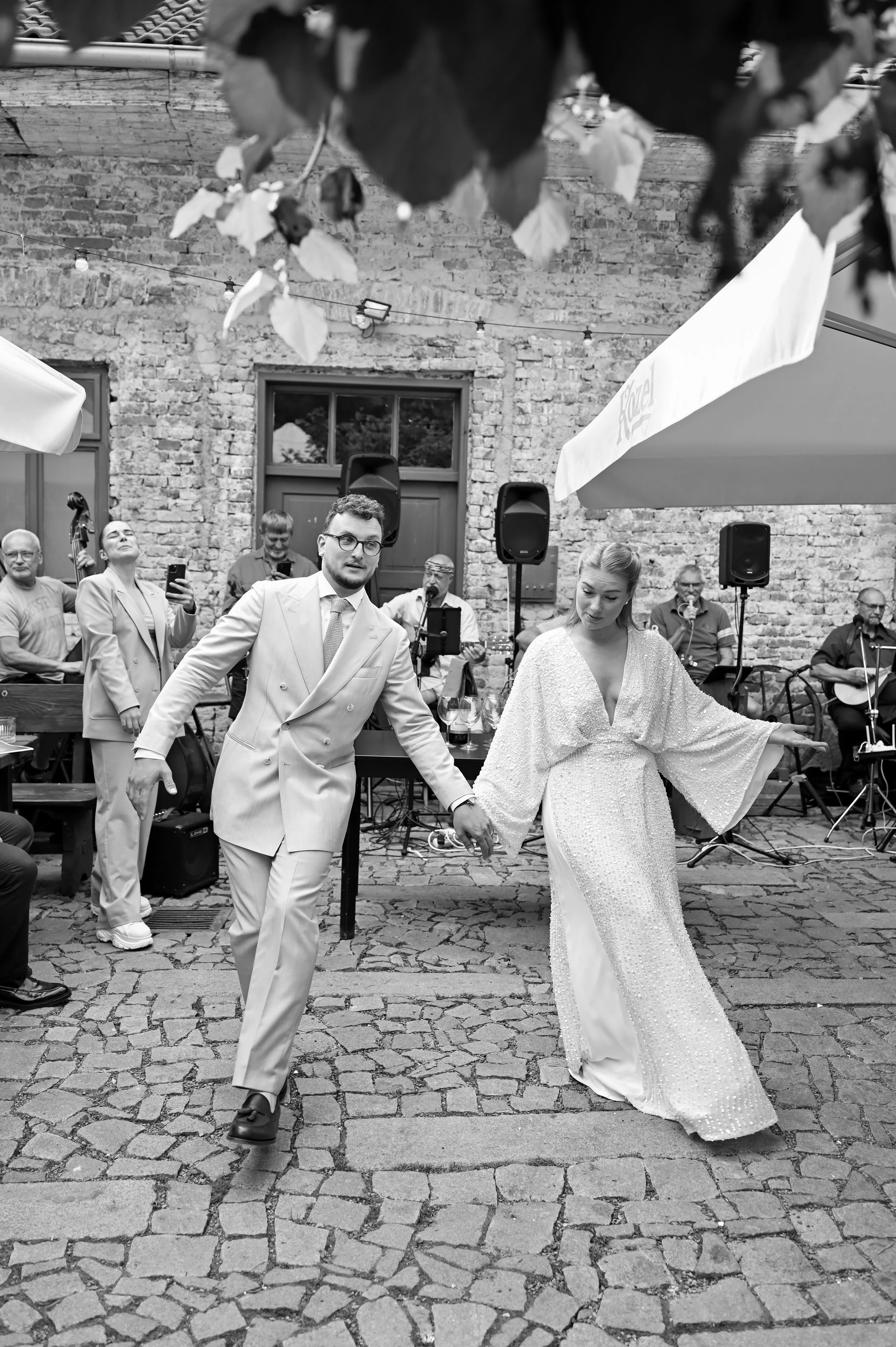 A black and white photo of a bride and groom dancing at an outdoor wedding reception, with a band playing in the background and guests watching.