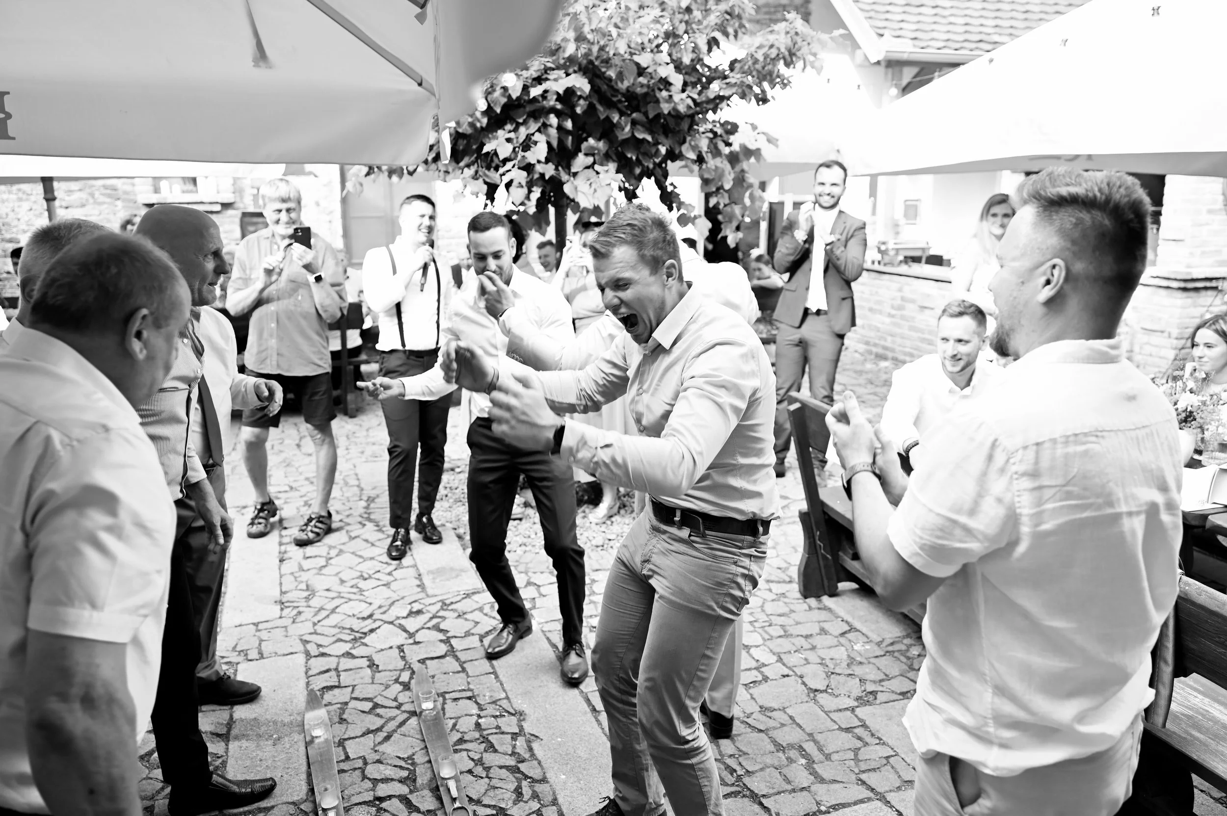 Group of men and women celebrating and dancing outdoors at a party, some clapping and taking pictures, with cobblestone ground and brick building in background.