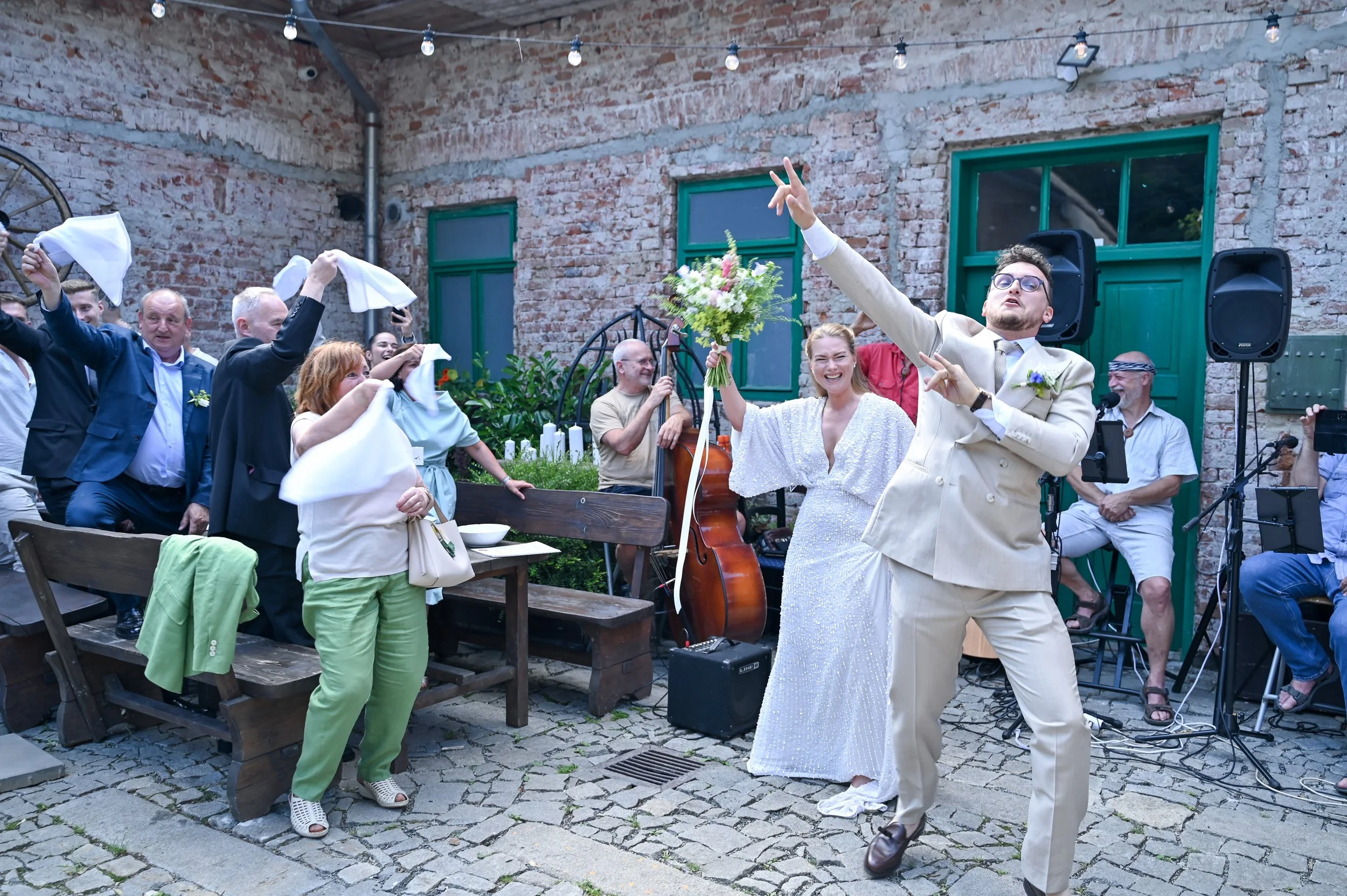 People celebrating at a wedding reception with the bride in a white dress smiling holding a bouquet, and a man in a beige suit dancing with arms outstretched, surrounded by guests clapping and cheering, in an outdoor courtyard with brick walls and st