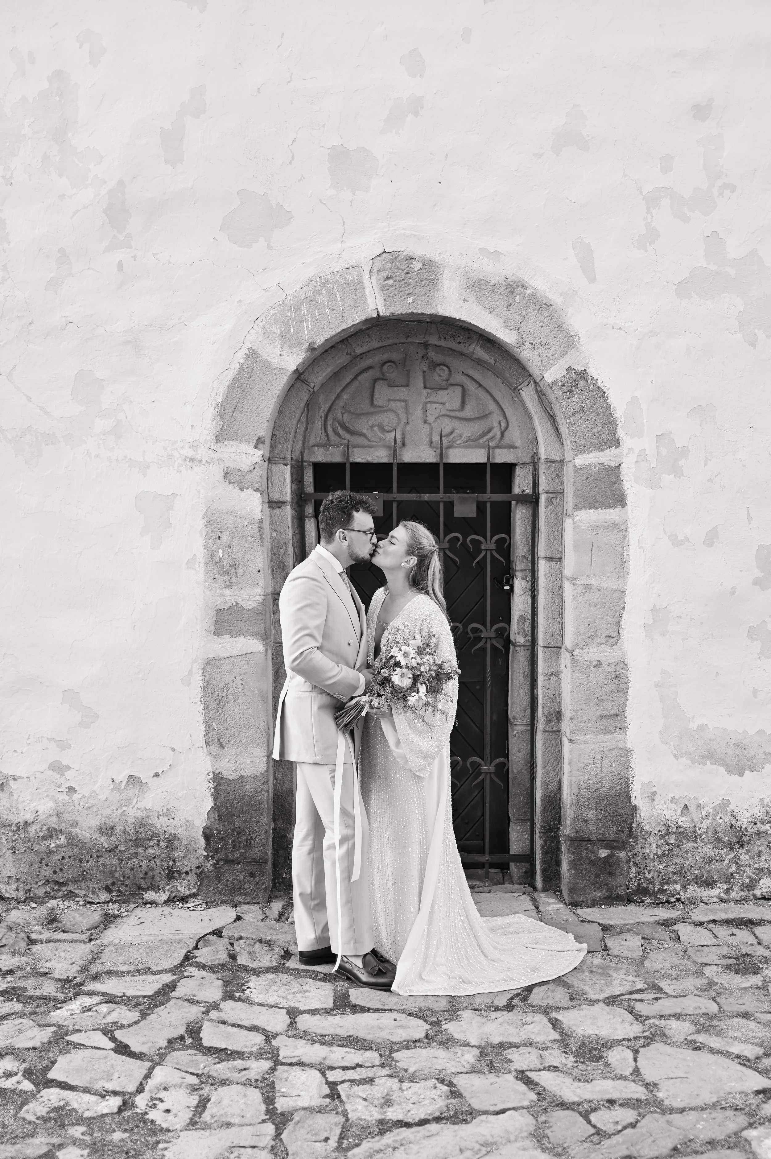 A black and white photo of a couple in wedding attire sharing a kiss in front of a stone archway with a barred door, on a cobblestone street.